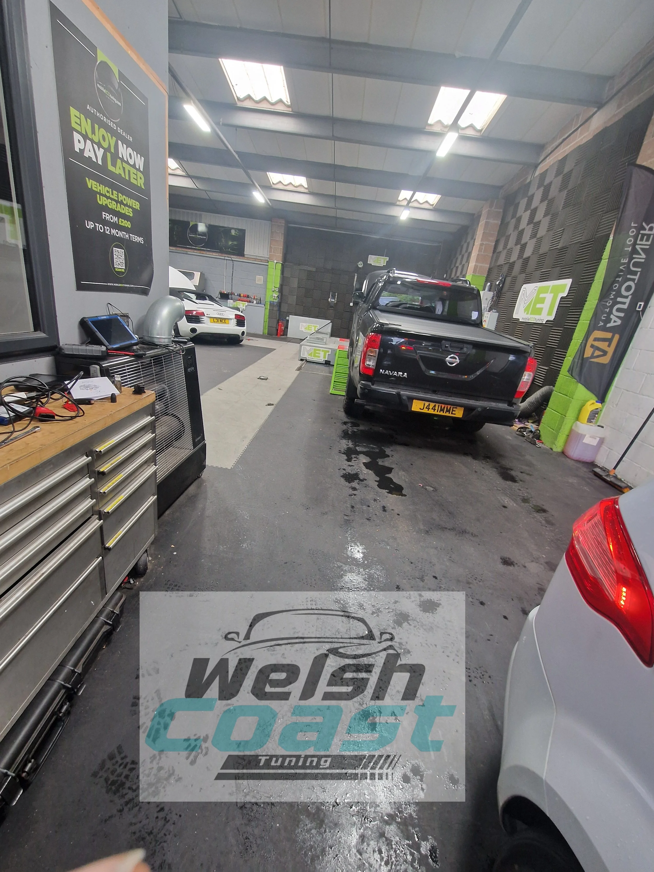 Inside an automotive workshop with cars being serviced, tools, and banners for vehicle upgrades and tuning.