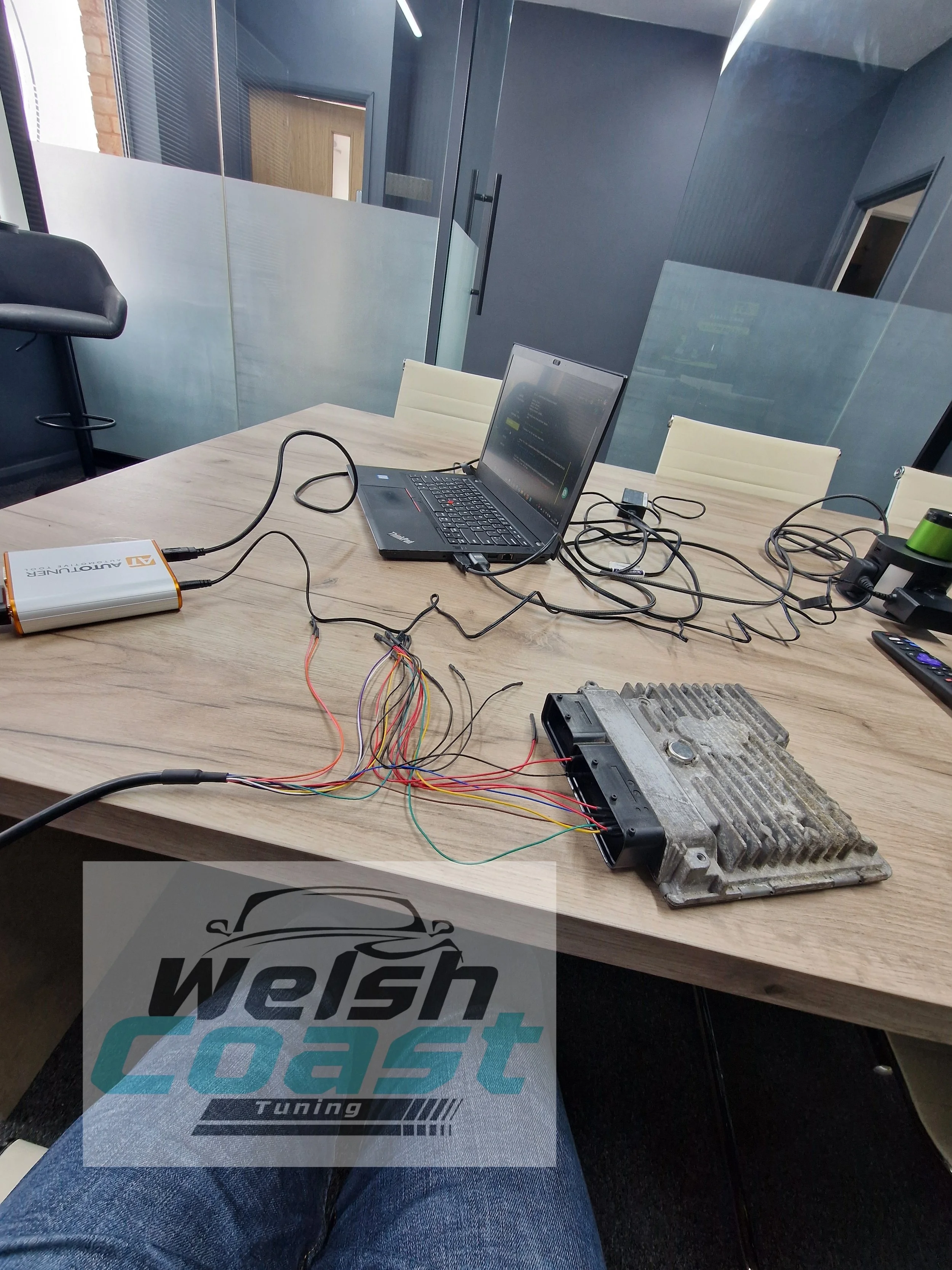 Table with wires, a laptop, an electronic device, and a heat sink in a conference room with glass walls.