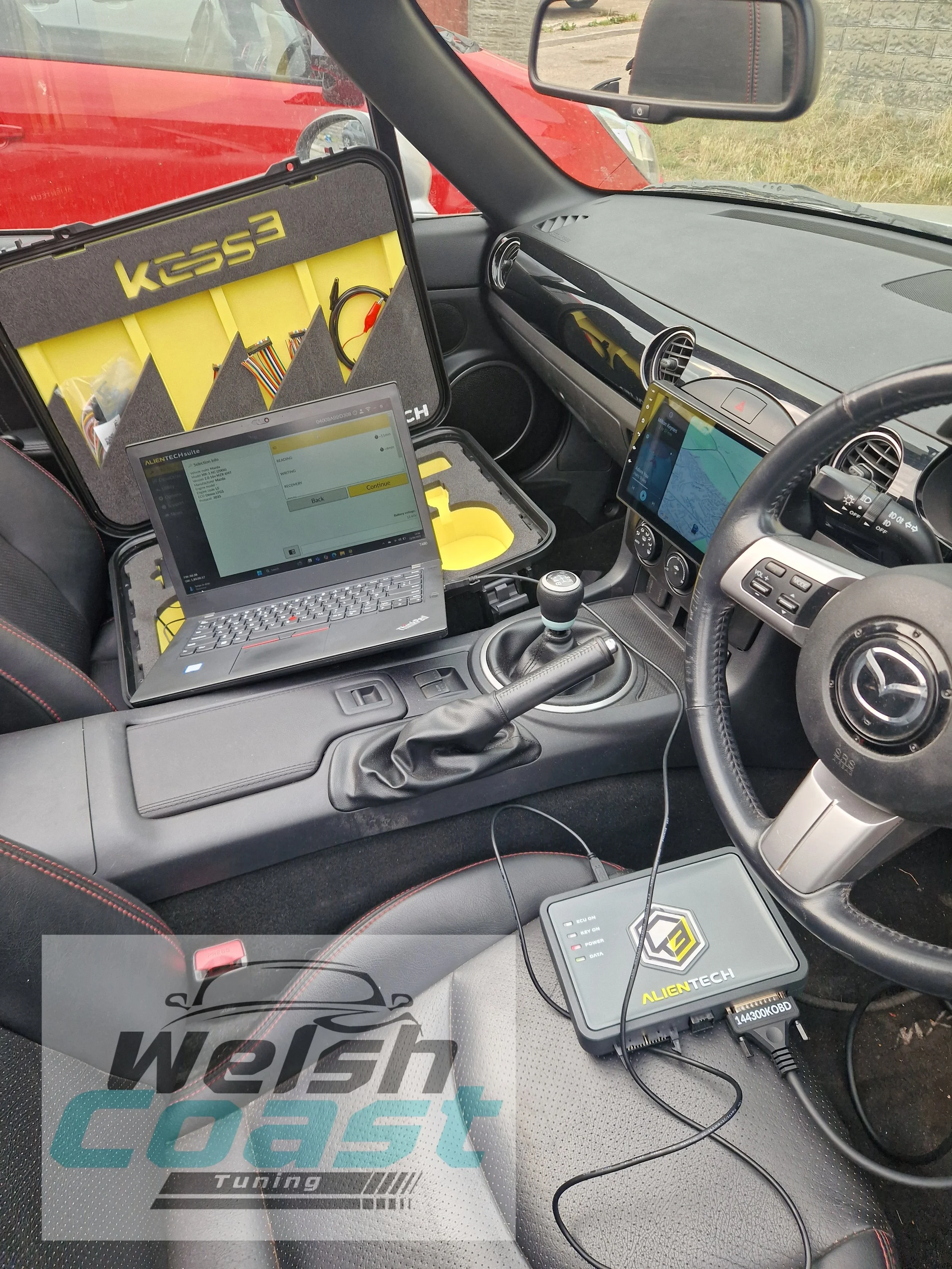 Inside a vehicle, a laptop and diagnostic tools are set up for vehicle tuning or diagnostics. There's a yellow and black kit box labeled 'KESS3,' and a device labeled 'ALIEN TECH' connected to the laptop with cables. The dashboard shows a digital screen with navigation, and there are other electronic devices nearby.