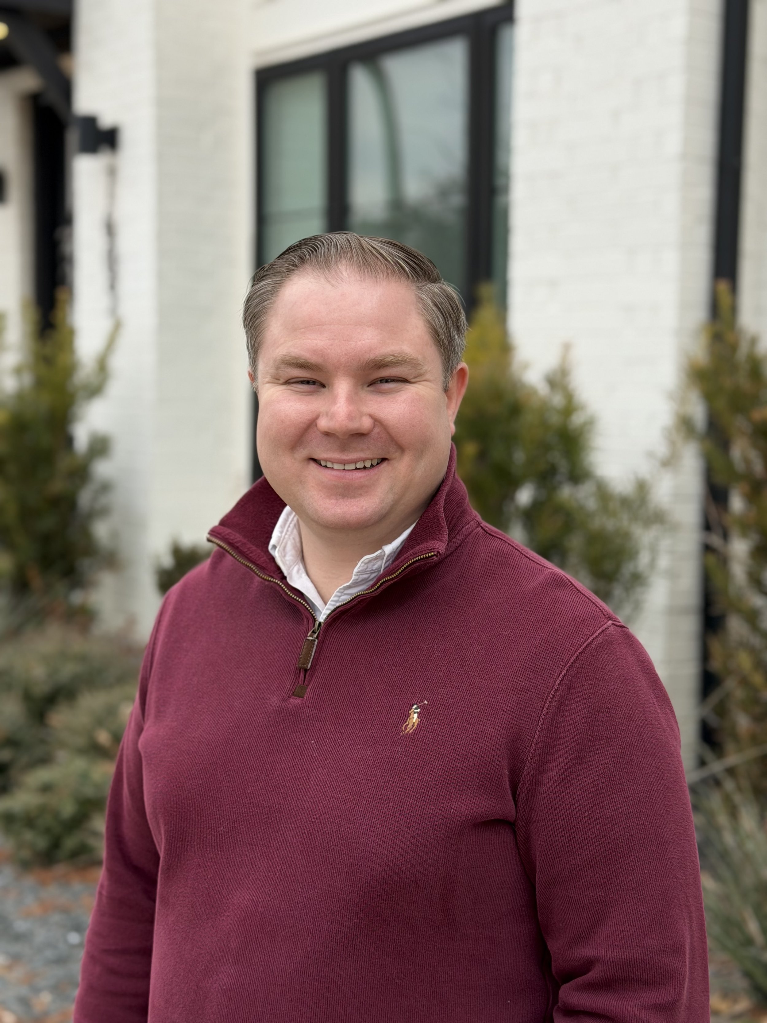 A man with short brown hair and a white shirt, smiling while sitting on a bed in a bedroom.