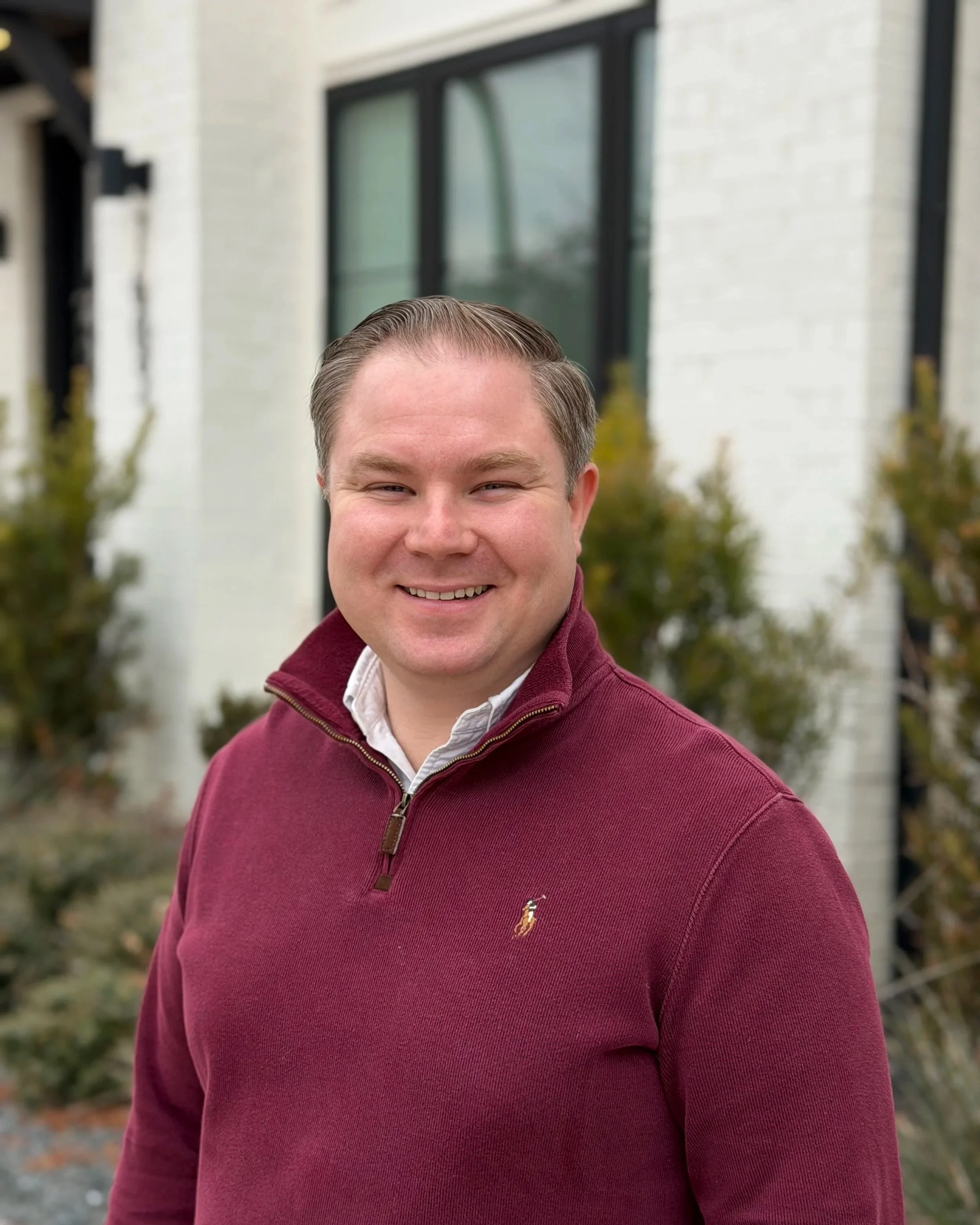 A smiling man in a burgundy quarter-zip sweater over a white collared shirt, standing outdoors in front of a white building with black trim and some foliage.