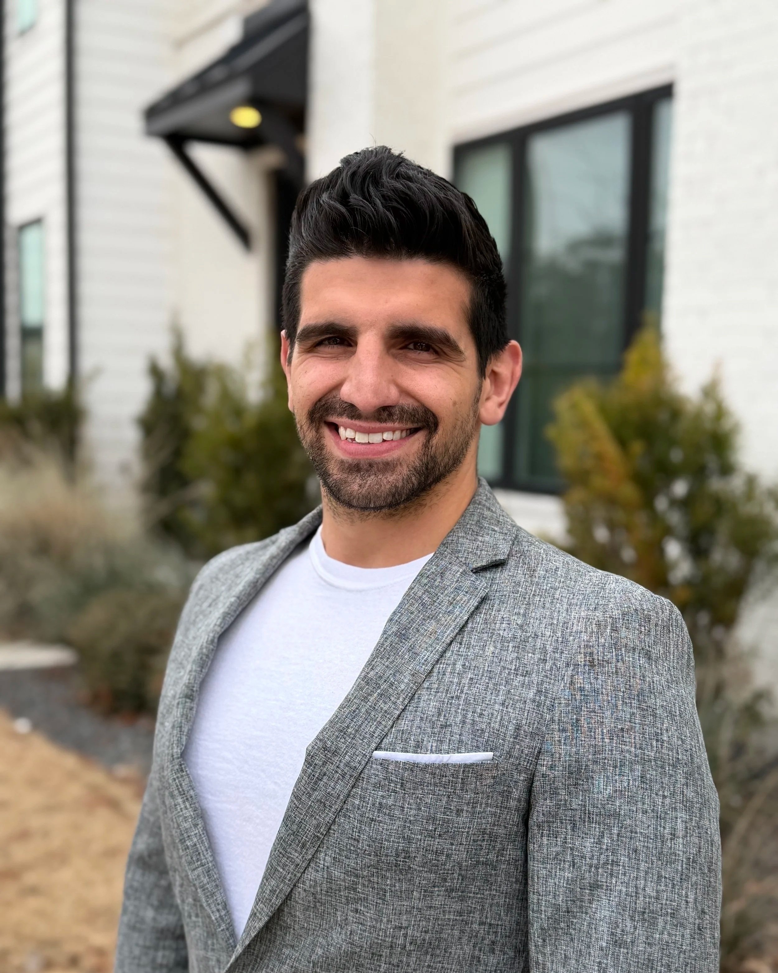 A man with dark hair and a beard smiling outdoors, wearing a gray blazer over a white shirt, with a house and bushes in the background.