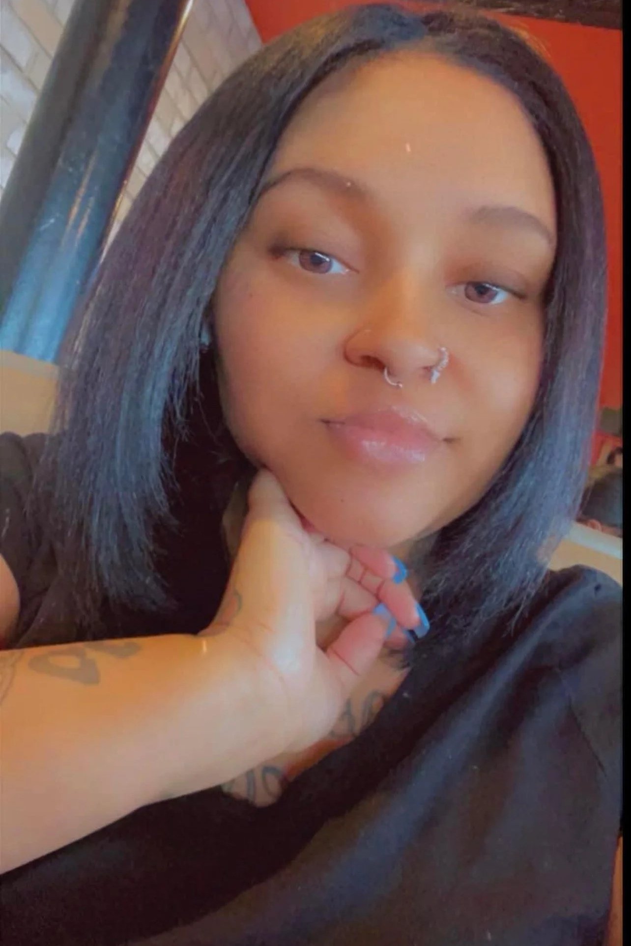 A young woman with straight black hair, light brown skin, and multiple facial piercings, resting her face on her hand, in a black shirt, sitting indoors with warm lighting.