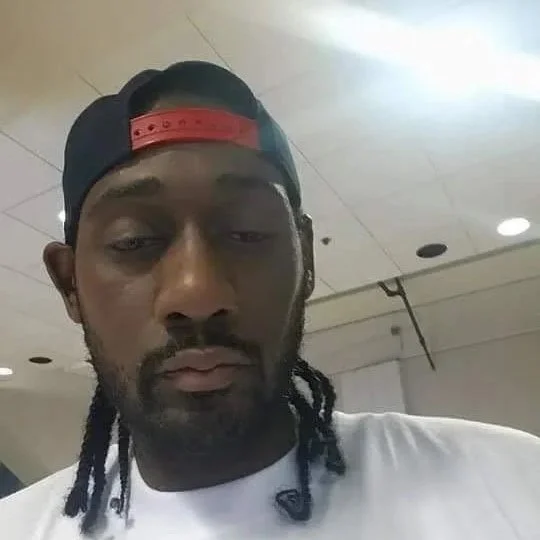 A man with braided hair wearing a backwards baseball cap, looking down with a neutral expression, in an indoor setting.