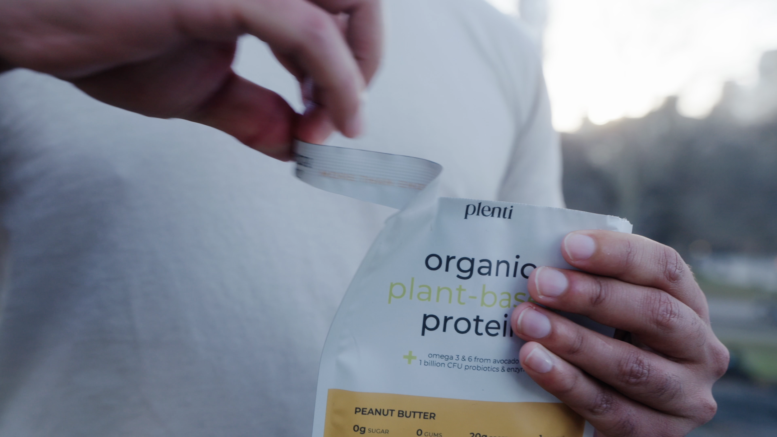 A person opening a package of organic plant-based protein powder labeled 'peanut butter' outside with a blurred background of trees and sky.