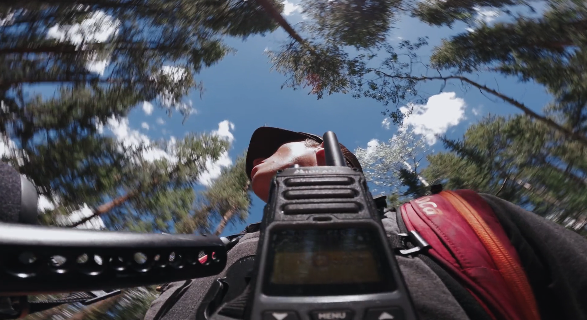 A person is taking a selfie from a low angle, looking up at the sky through tall trees with a few clouds, holding a camera or device, and wearing a hat and backpack.