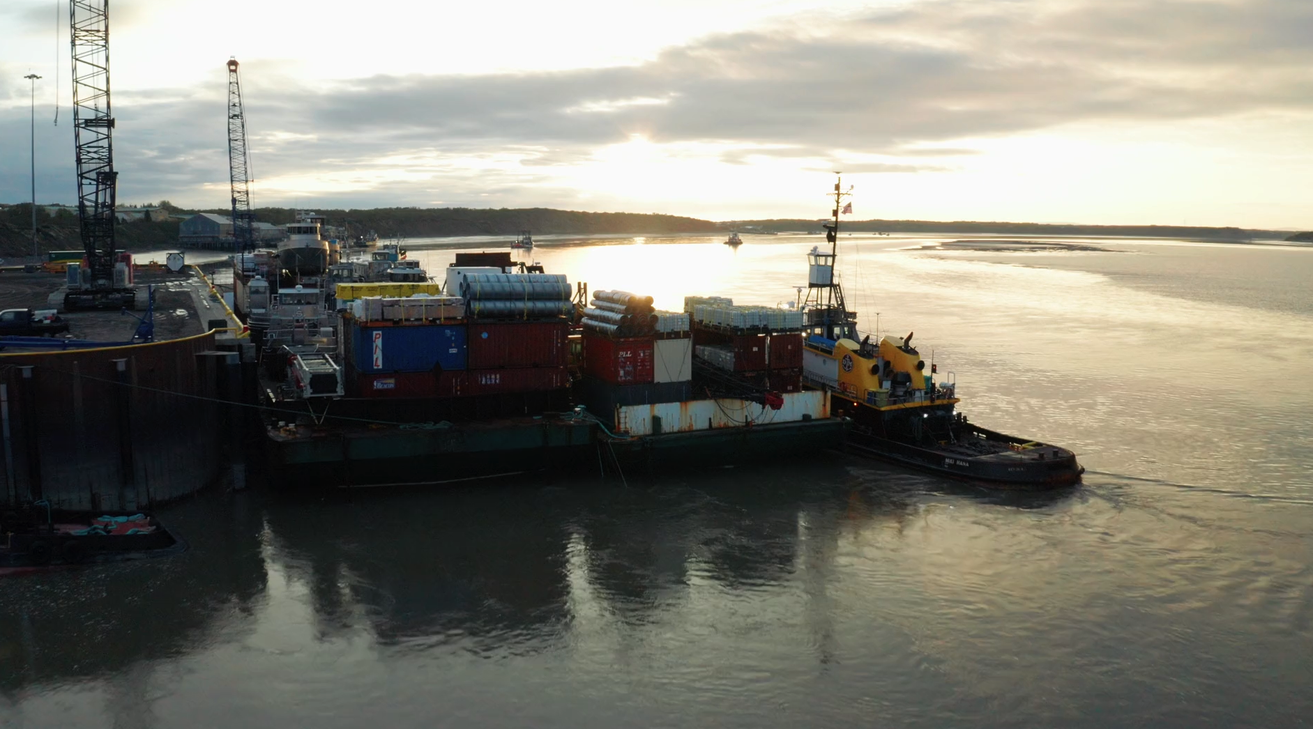 A barge carrying shipping containers and equipment being guided by a tugboat in a river at sunset.