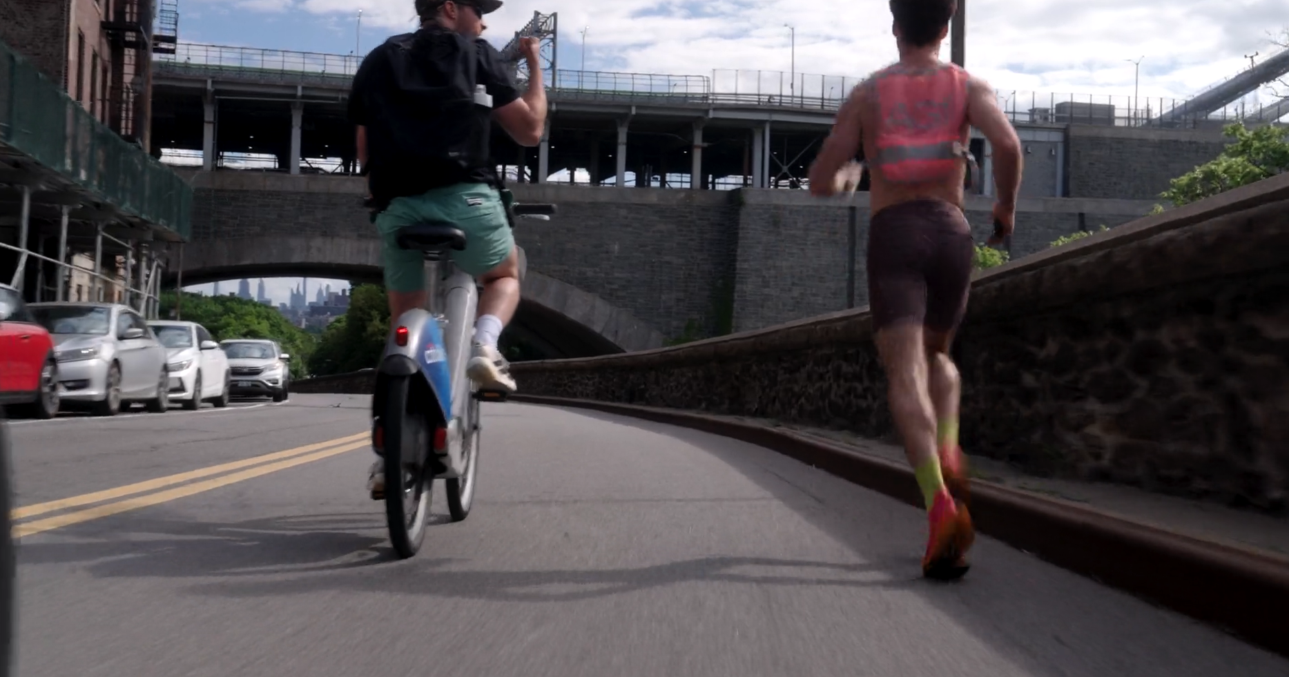 A cyclist and a runner on a city street under a bridge with cars parked alongside and skyscrapers in the background.