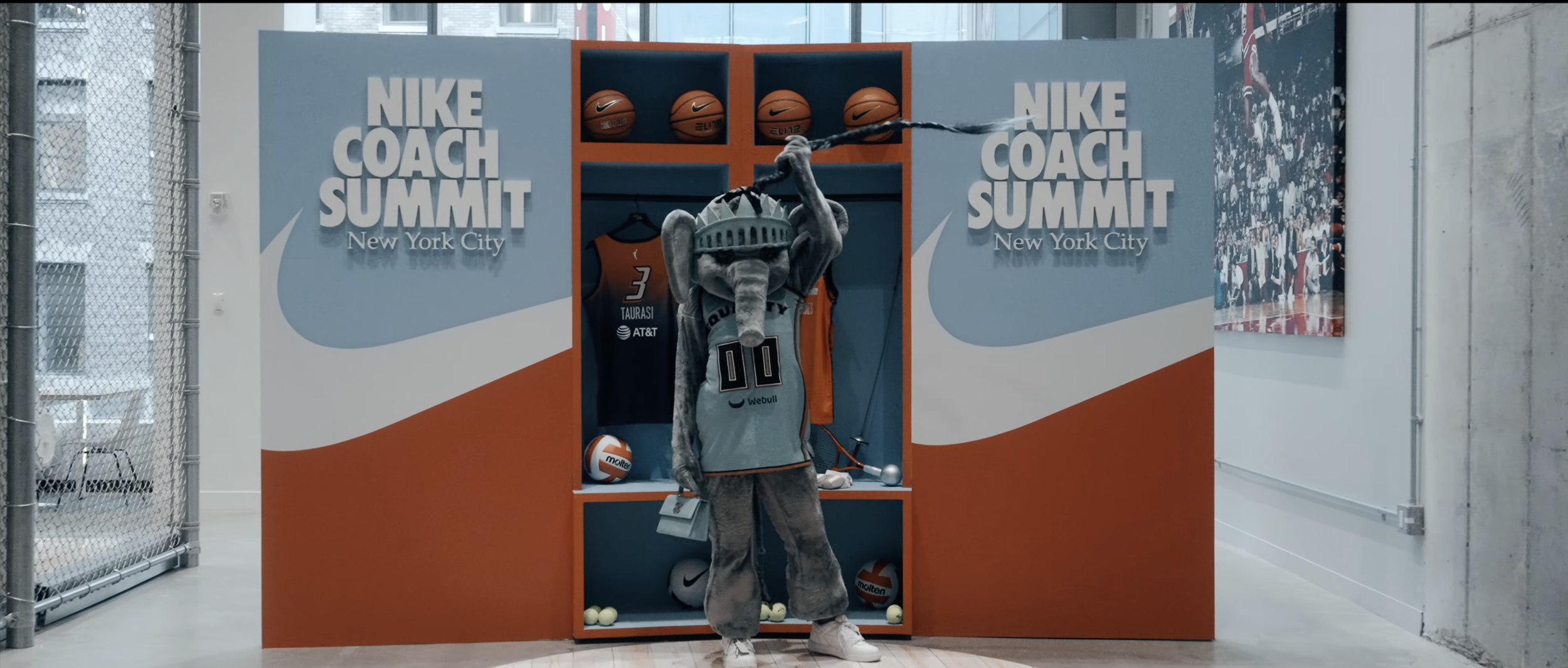 A Nike basketball-themed display featuring a mascot dressed as an elephant wearing a New York Knicks jersey, standing in front of lockers with basketballs and sports equipment, at the Nike Coach Summit in New York City.