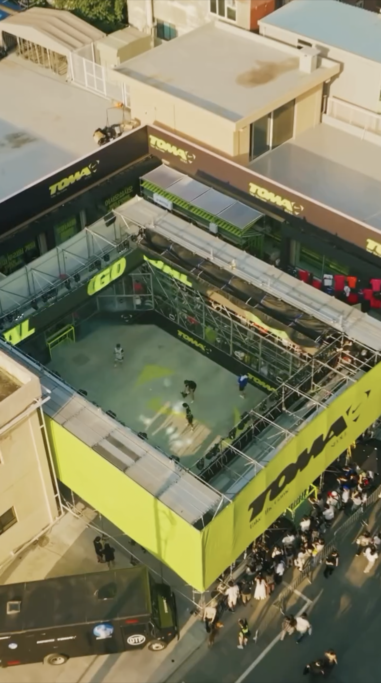 A rooftop miniature soccer field with players, a crowd below, and surrounding retail stores with prominent yellow and black branding.