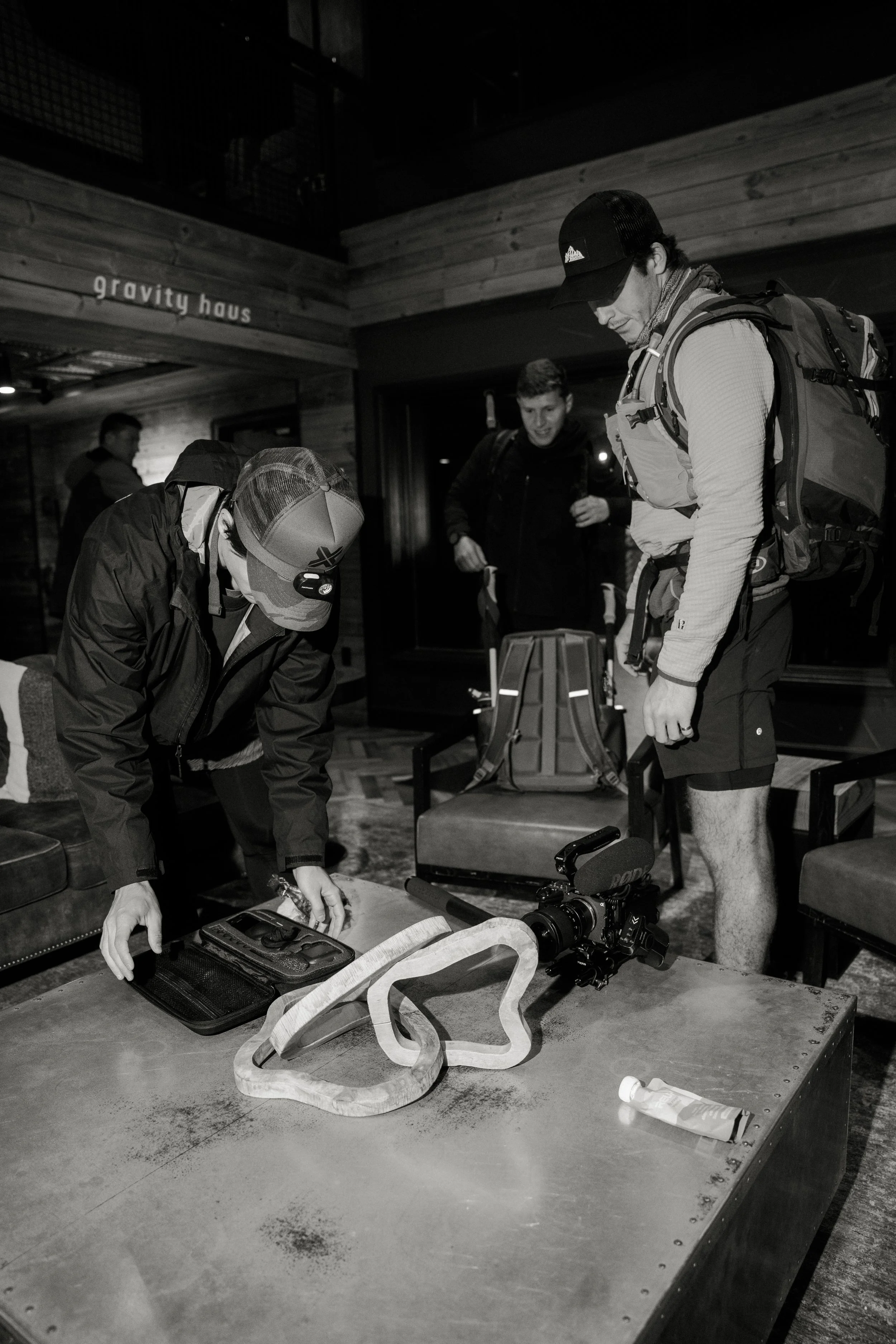 Two men preparing camera equipment and gear for filming indoors, with one man bending over a table, and the other standing and looking down. A person in the background is holding a backpack. The setting appears to be a rustic, wooden interior with a sign that reads 'gravity haus' on the wall.