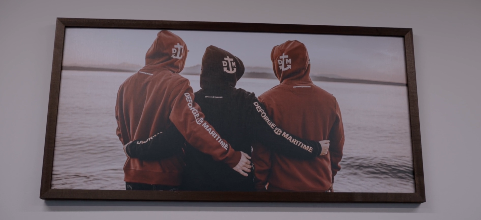 Three people in hooded jackets standing by a lake, with arms around each other's shoulders, facing away from the camera.