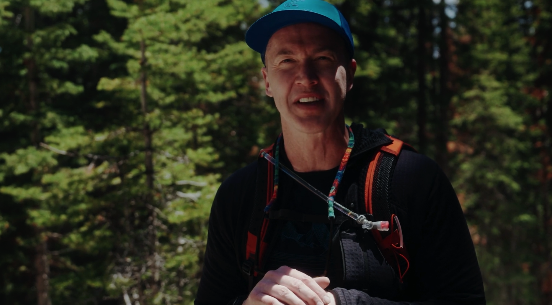 A man wearing a blue cap and black athletic clothing with an orange hydration pack, outdoors in a wooded area.