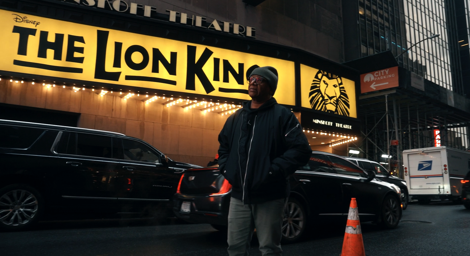 A person standing on a city street at night in front of a large illuminated sign for "The Lion King" musical at the Minskoff Theatre, with a car and a postal truck in the background.