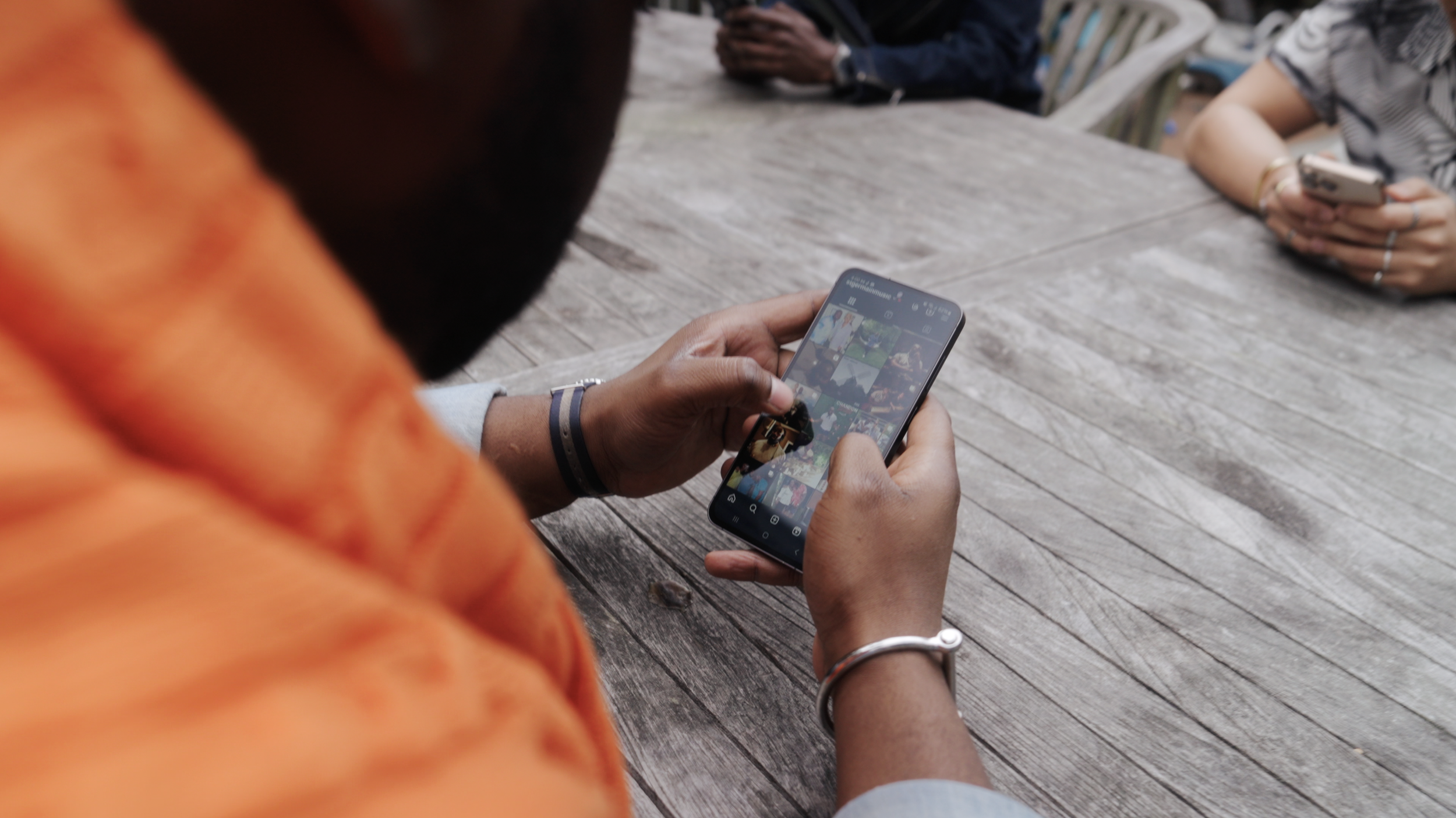 Person holding a smartphone over a wooden table, with other people sitting at the table using their phones in the background.