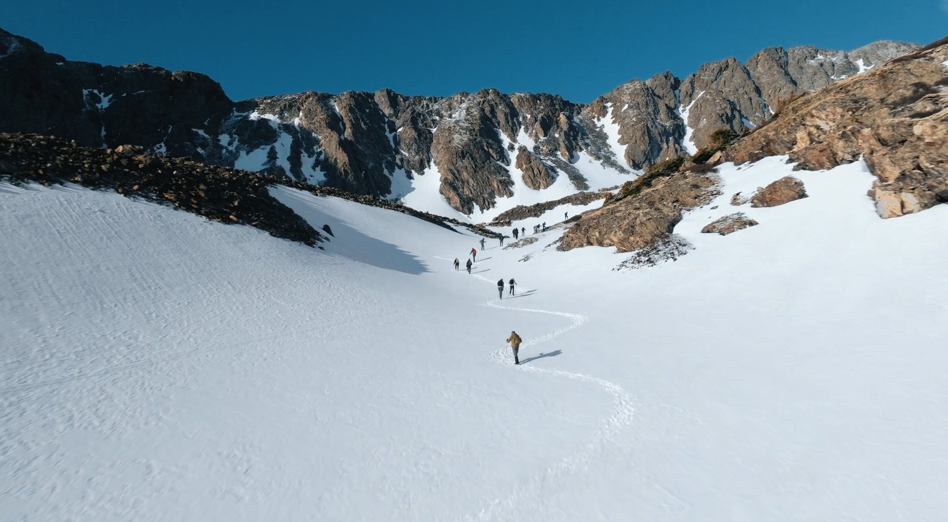 A group of hikers trekking through a snowy mountain landscape with rocky peaks and a clear blue sky.