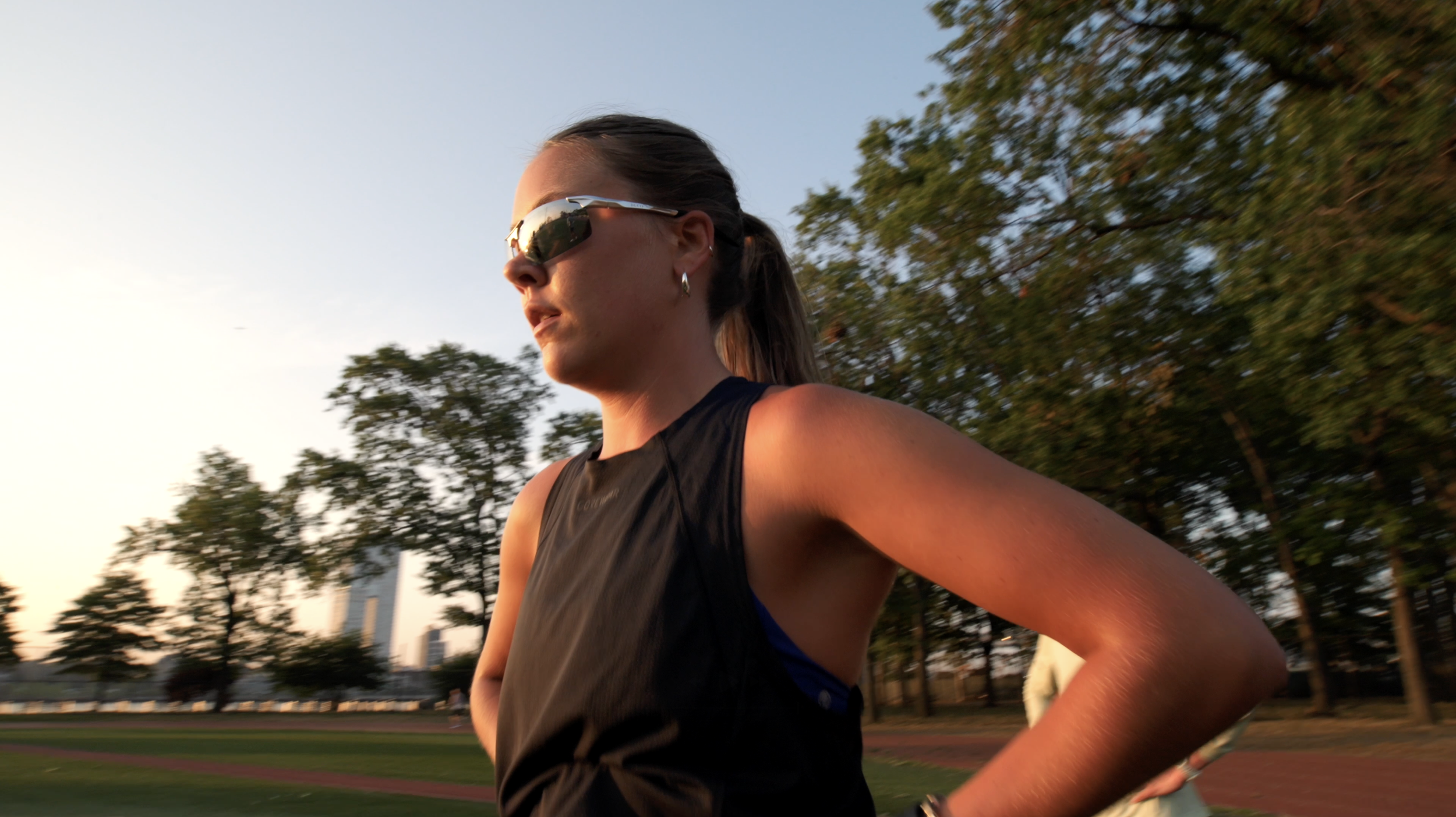 A woman running outdoors during sunset, wearing sunglasses and a black athletic shirt, with trees and a cityscape in the background.