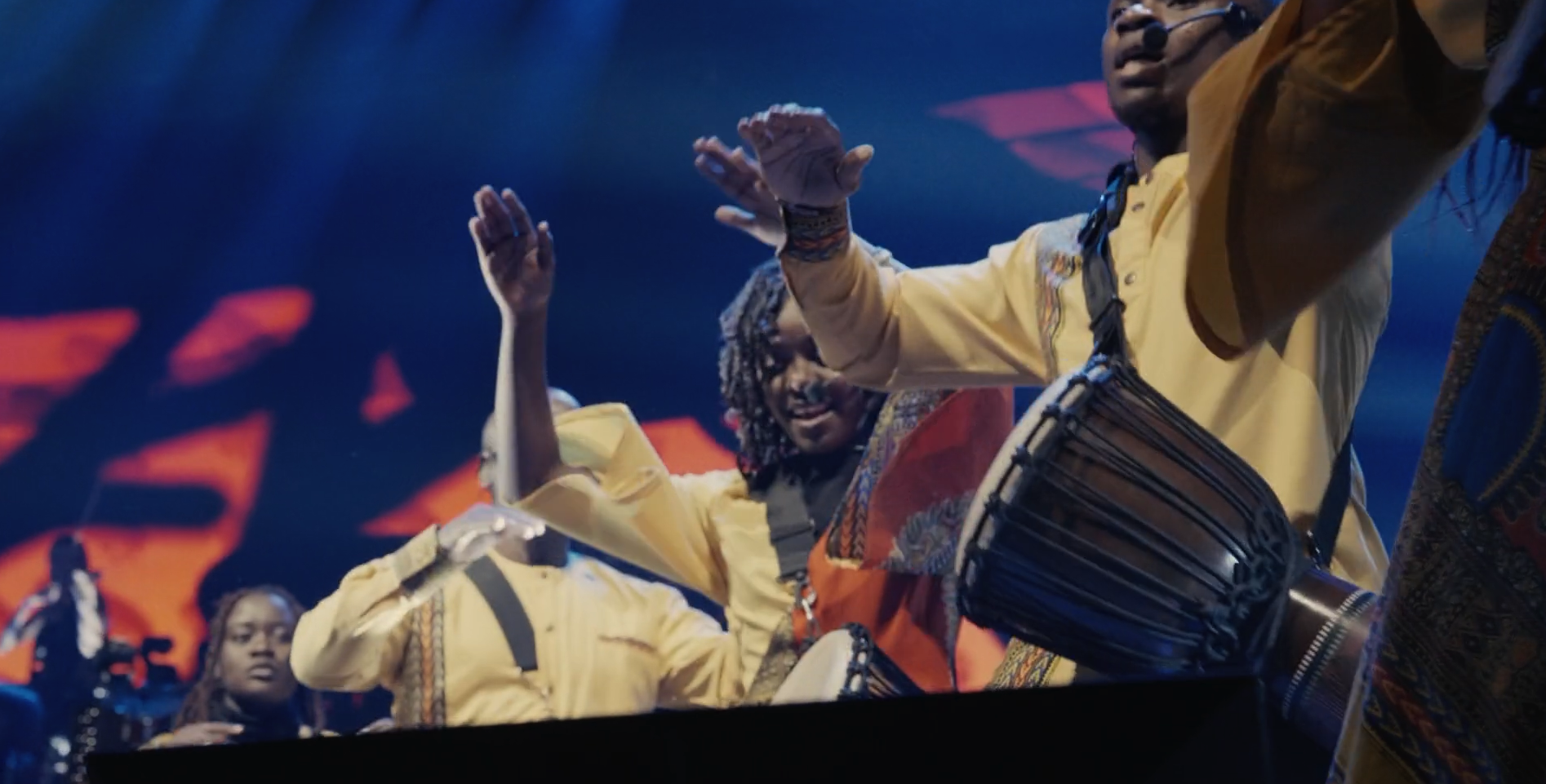 Musicians in traditional attire playing drums and dancing on stage with a dark background and stage lighting.