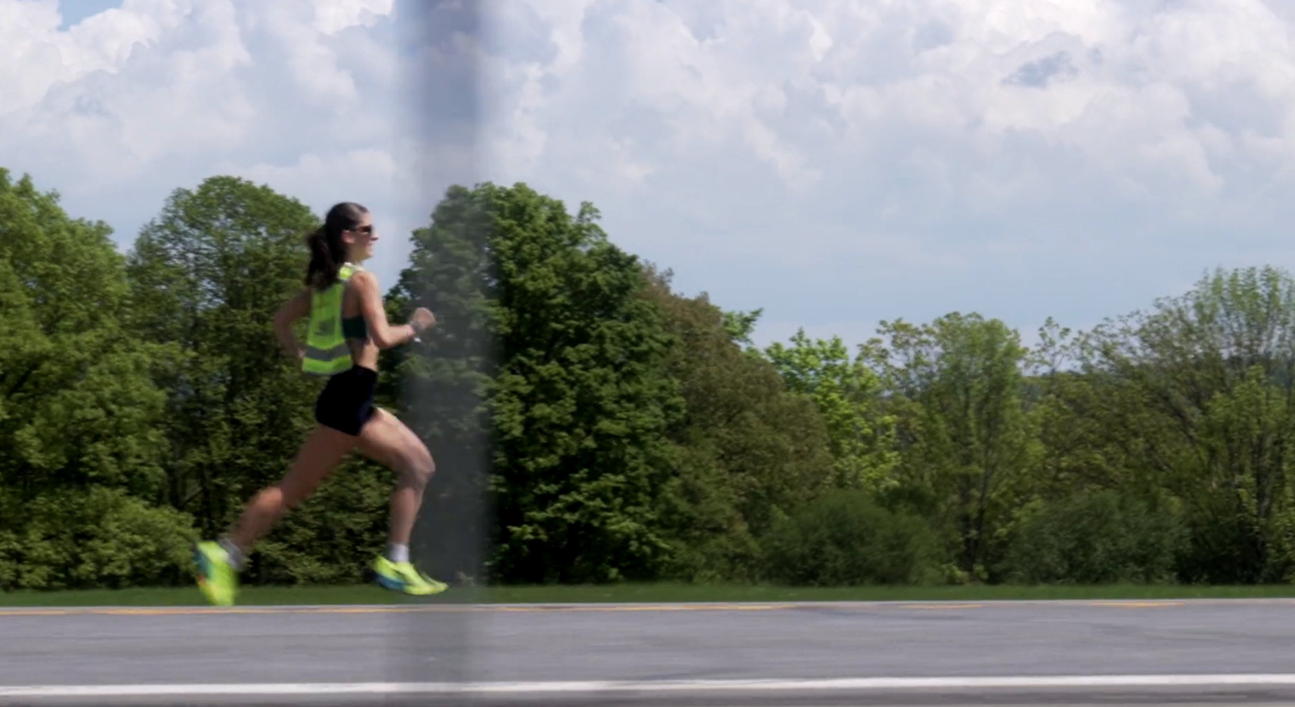 A woman running outdoors on a sunny day, wearing a bright yellow vest, black shorts, and running shoes.