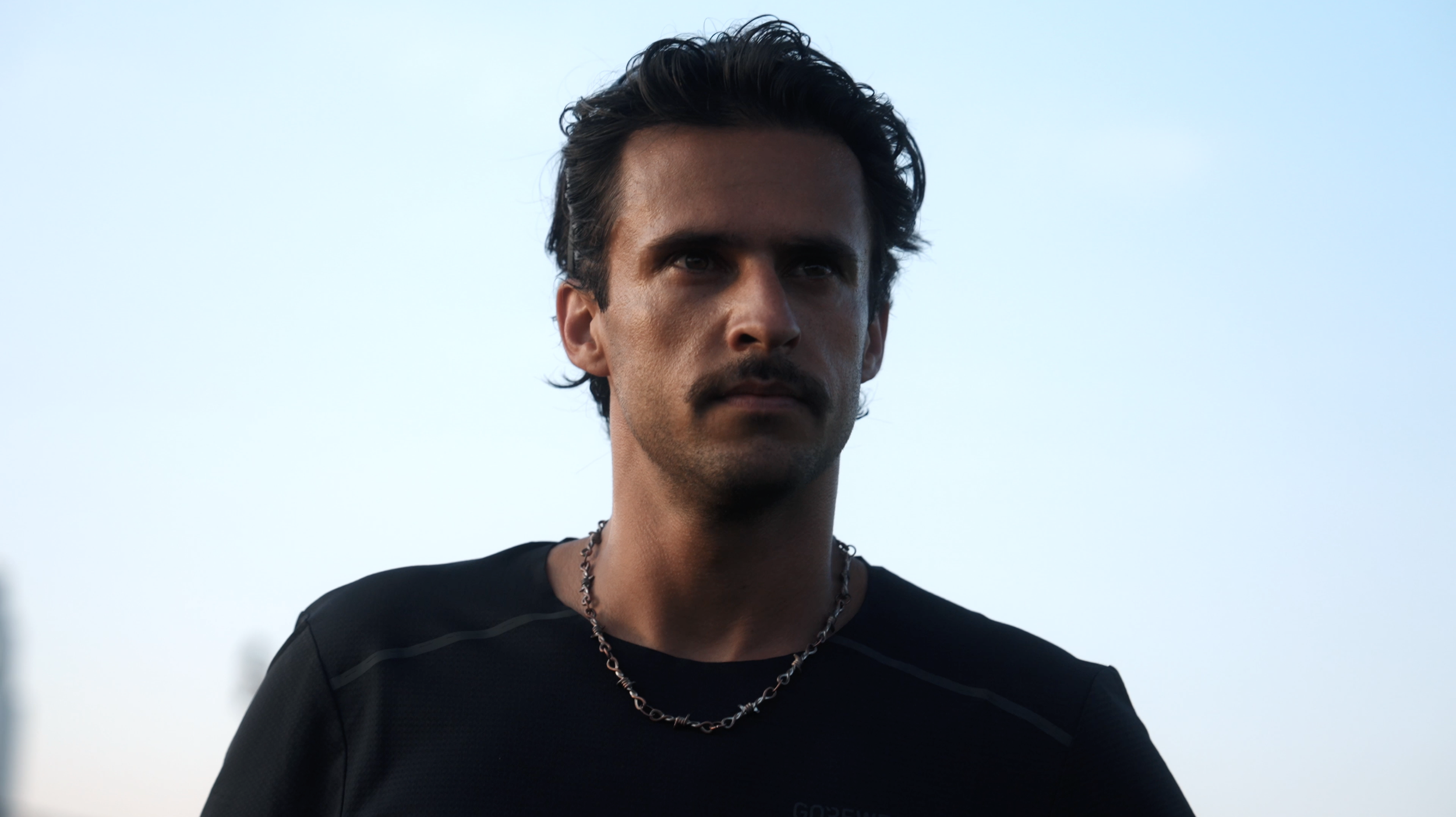 A man with dark hair, mustache, and a serious expression standing outdoors against a clear blue sky, wearing a black shirt and a silver chain necklace.