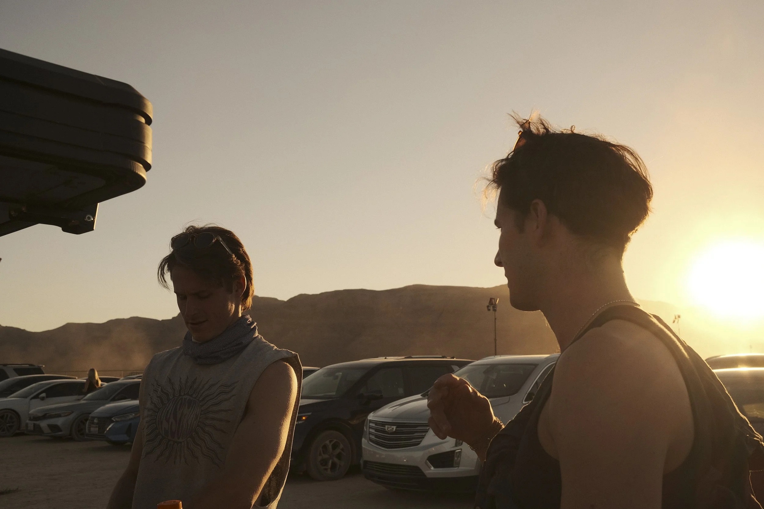 Two young men are standing in a parking lot at sunset, with parked cars and mountains in the background.