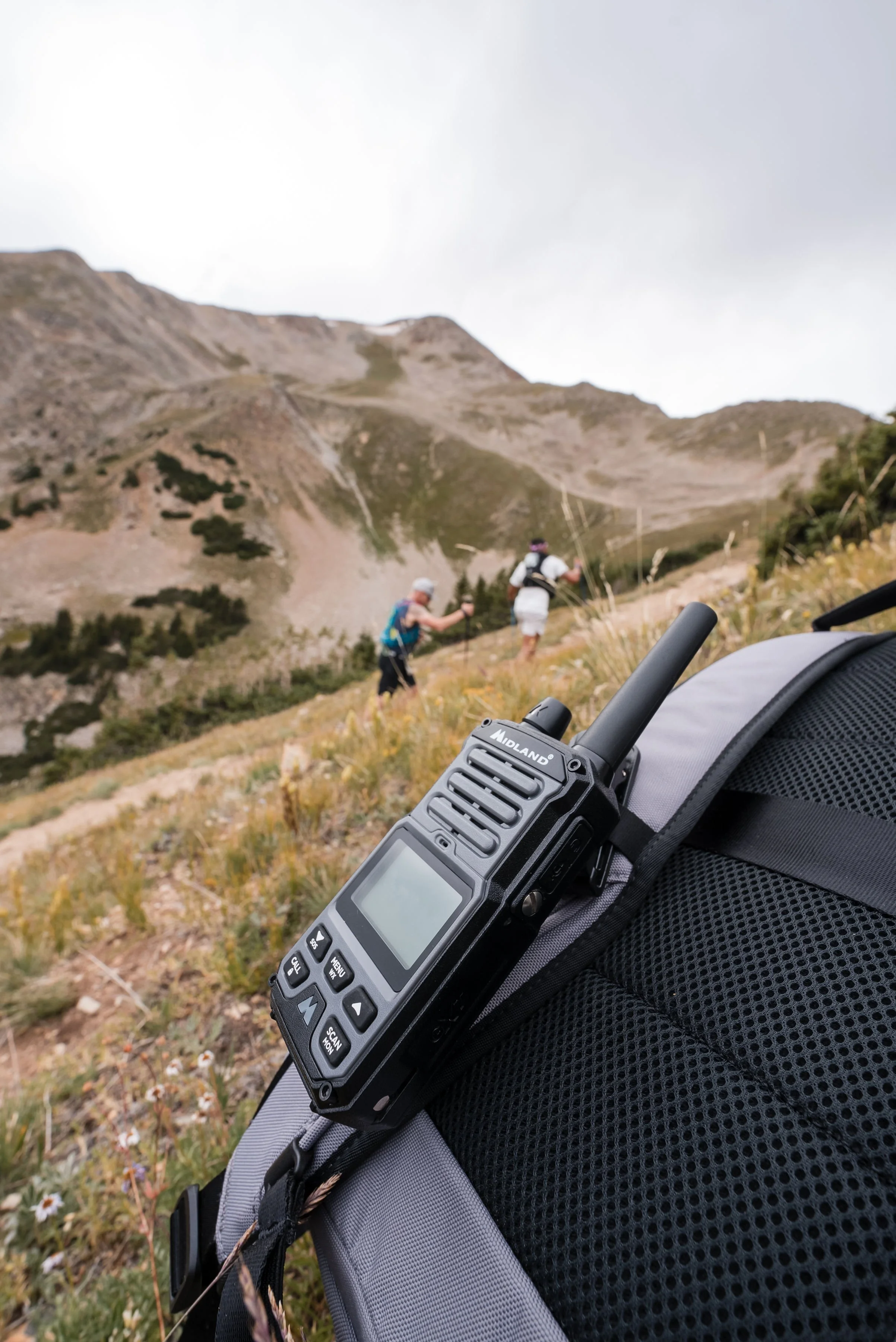 A handheld radio on a backpack with two hikers trekking up a mountain trail in the background.
