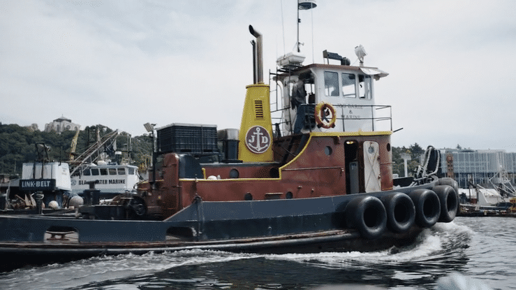 A tugboat moving across the water at a harbor with other boats and shoreline in the background.