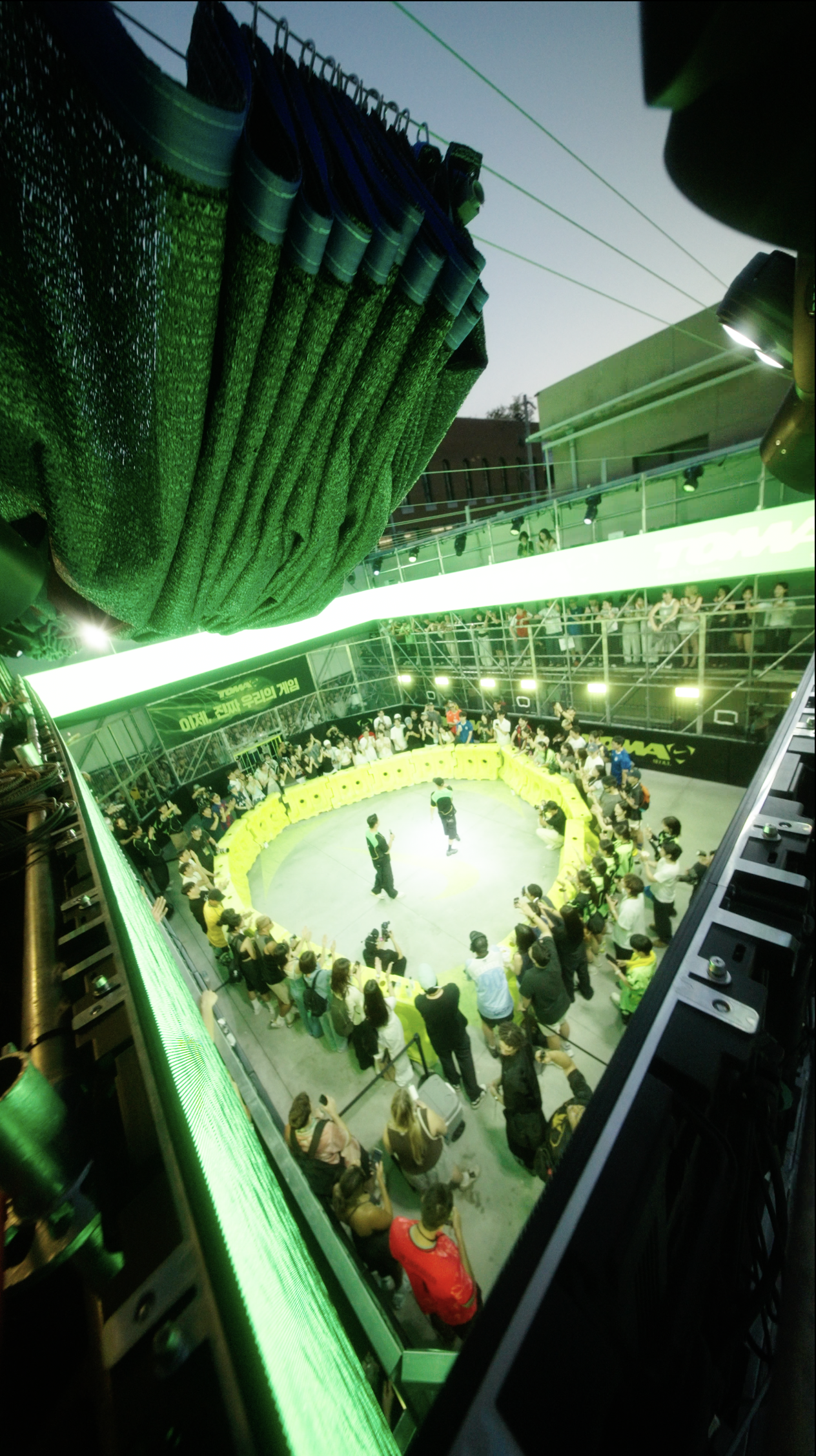 An aerial view of a large indoor event or competition with a circular stage, surrounded by spectators and photographers. There are green and black banners, with some people standing inside the stage area and others watching from the surrounding seats.