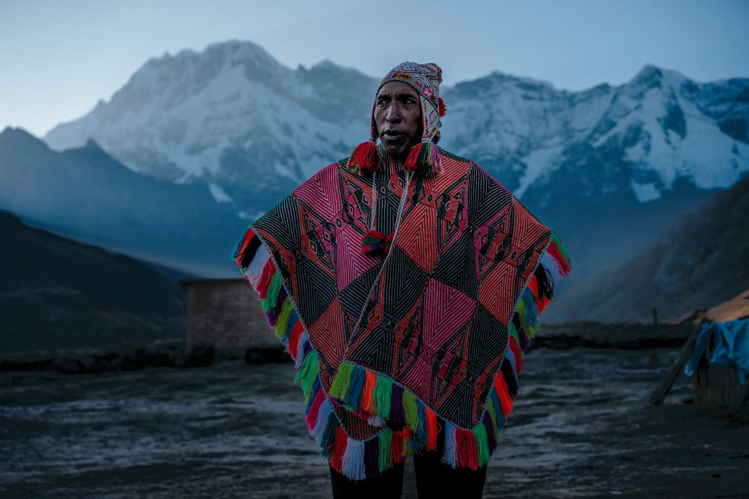 Man wearing traditional colorful woven poncho and hat standing outdoors in mountainous landscape with snow-capped peaks at dusk.