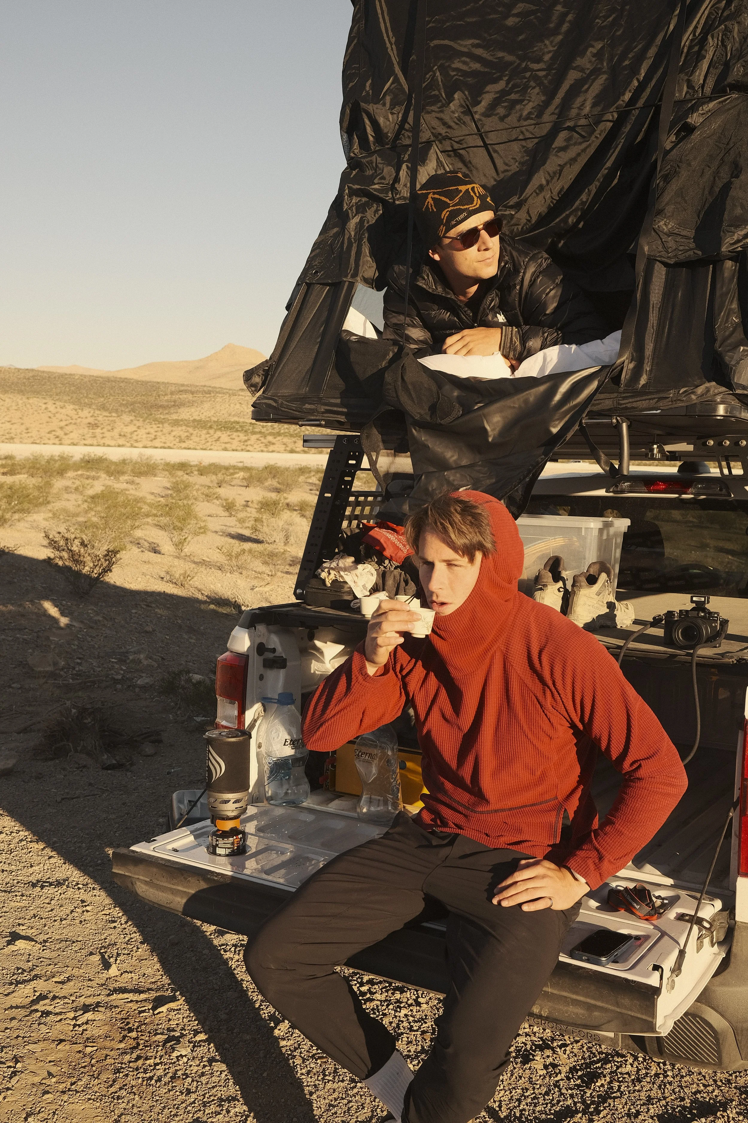 Two young men with short hair and casual outdoor clothing, one lying inside a black, elevated tent and the other sitting on the back of a pickup truck, drinking from a cup in a desert landscape with mountains in the background during sunset.