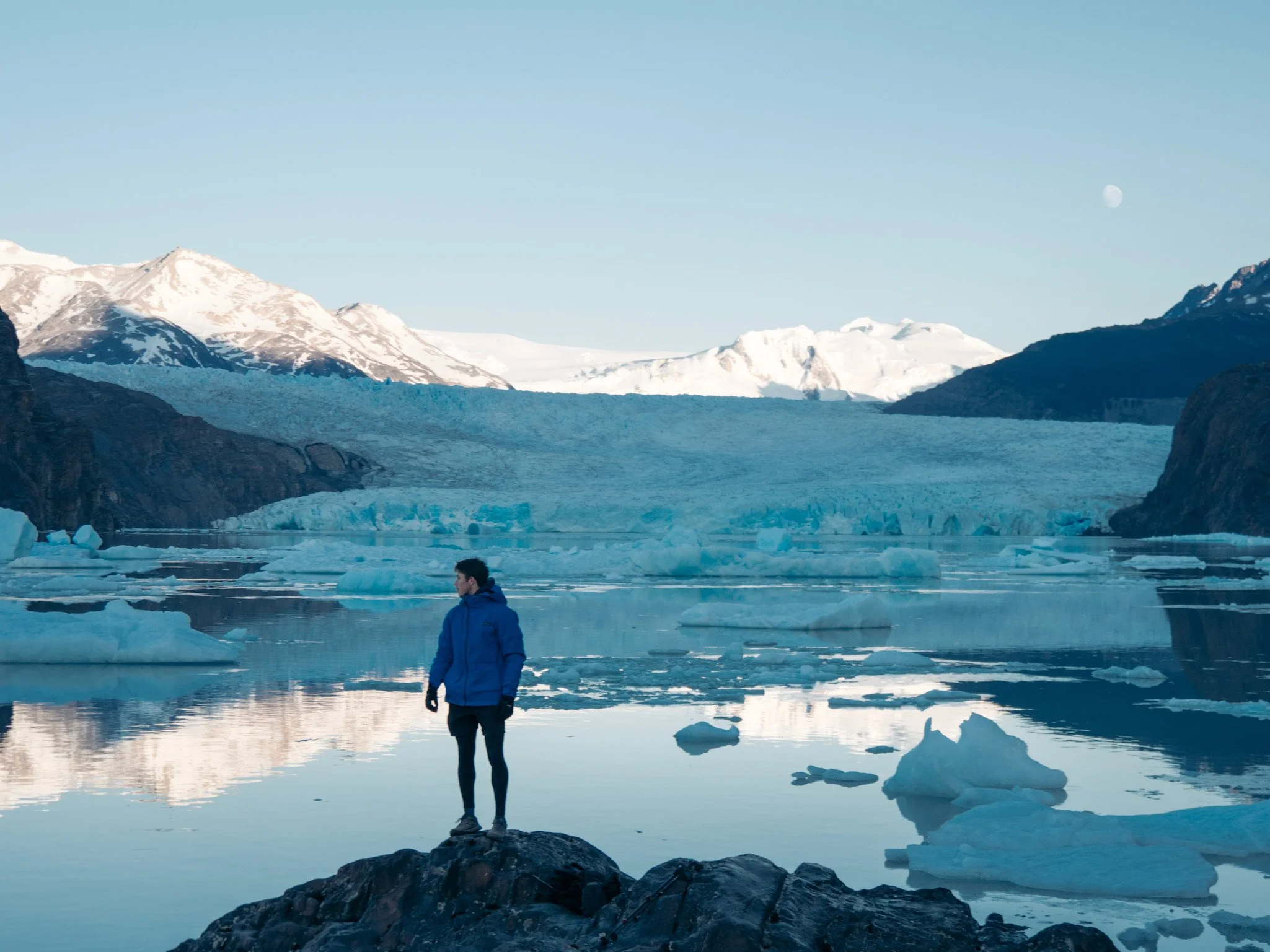 A person in a blue jacket and black pants standing on a rock near icy water, with icebergs, glaciers, snow-capped mountains, and a visible moon in the sky in the background.