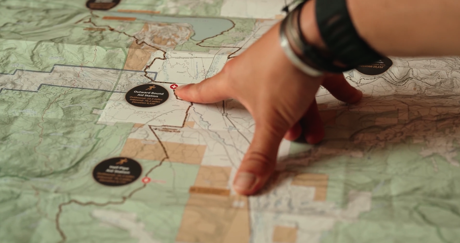 Close-up of a person's hand pointing at a map, with a focus on an area labeled 'Outward Bound Station,' indicating a trail or location in a natural setting.