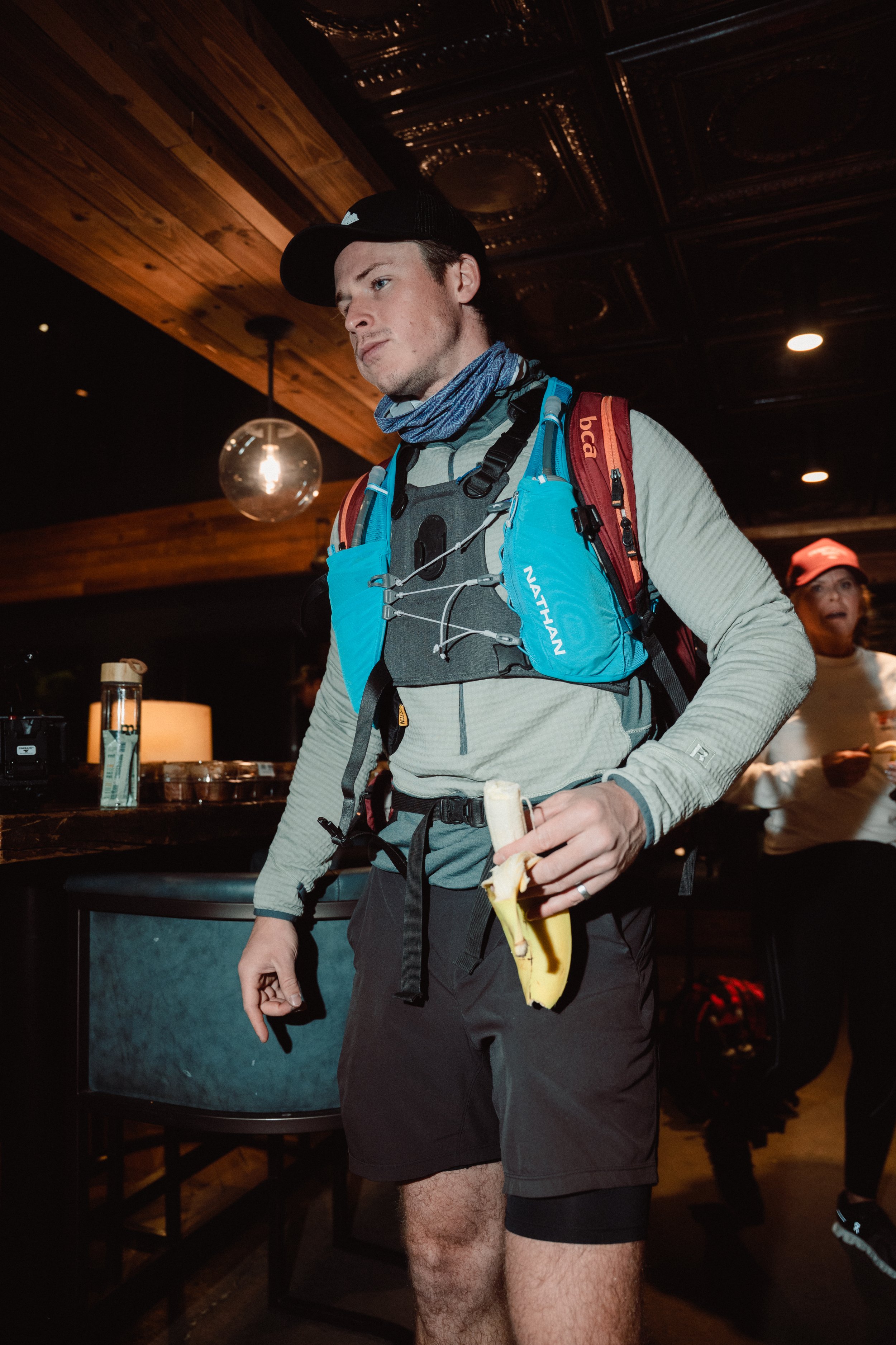 A young man in outdoor gear, holding a banana and a banana-flavored snack, standing indoors under a wood-paneled ceiling with a hanging light fixture, with a woman in a red hat and white shirt in the background.