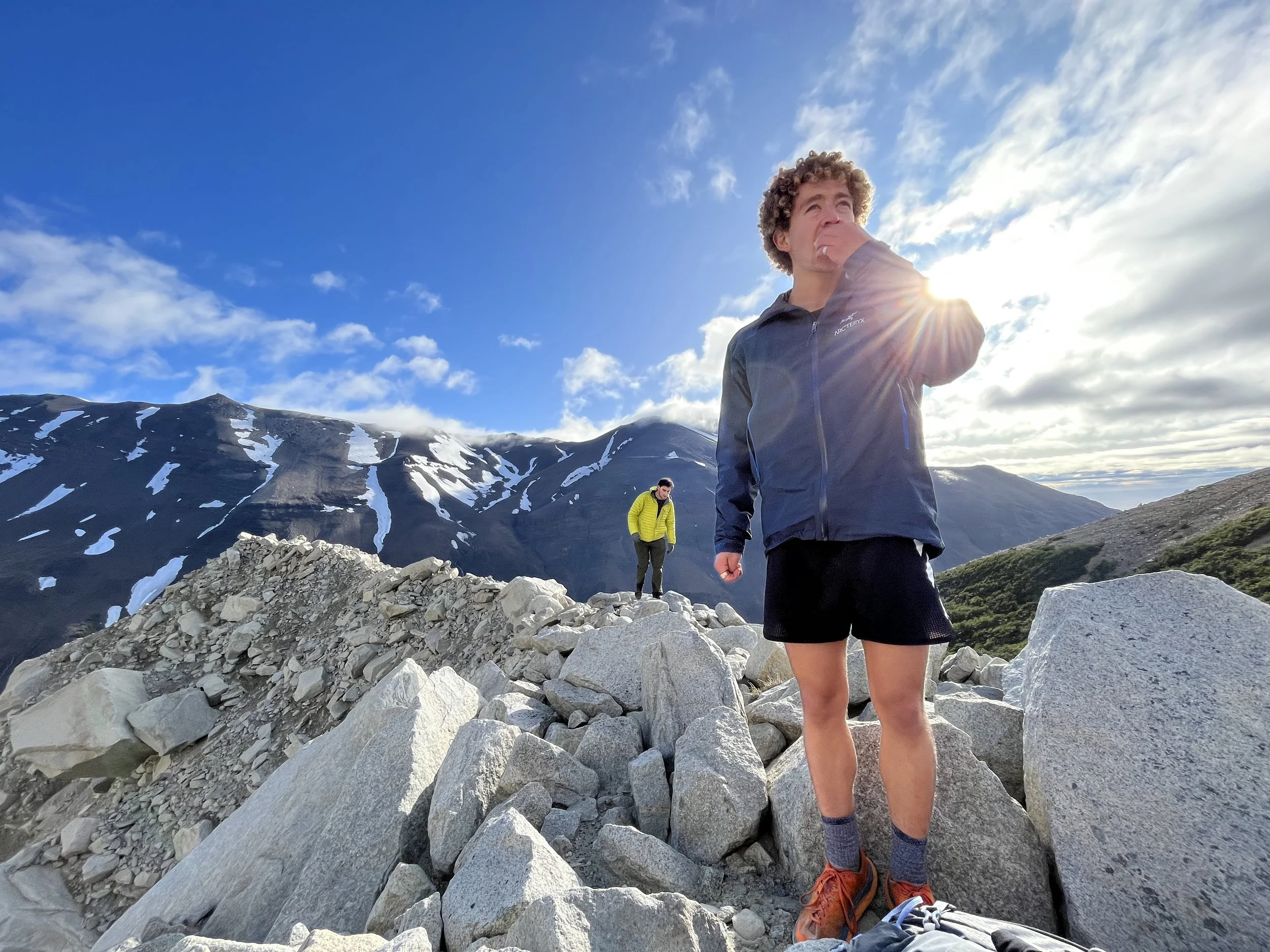 Two hikers standing on rocky terrain in a mountainous area, with snow-capped peaks and a partly cloudy sky in the background.