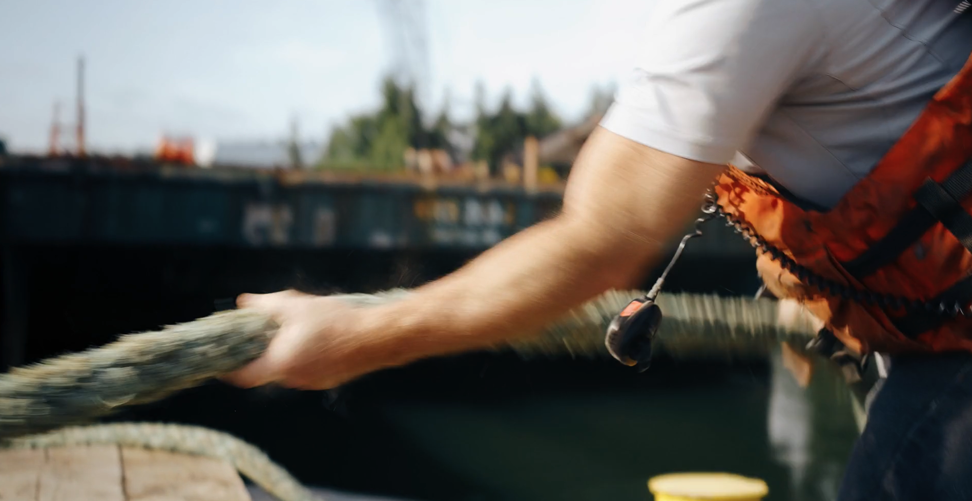 A person pulling a thick, brown rope on a boat, wearing a white shirt and orange life vest, with a marine environment in the background.