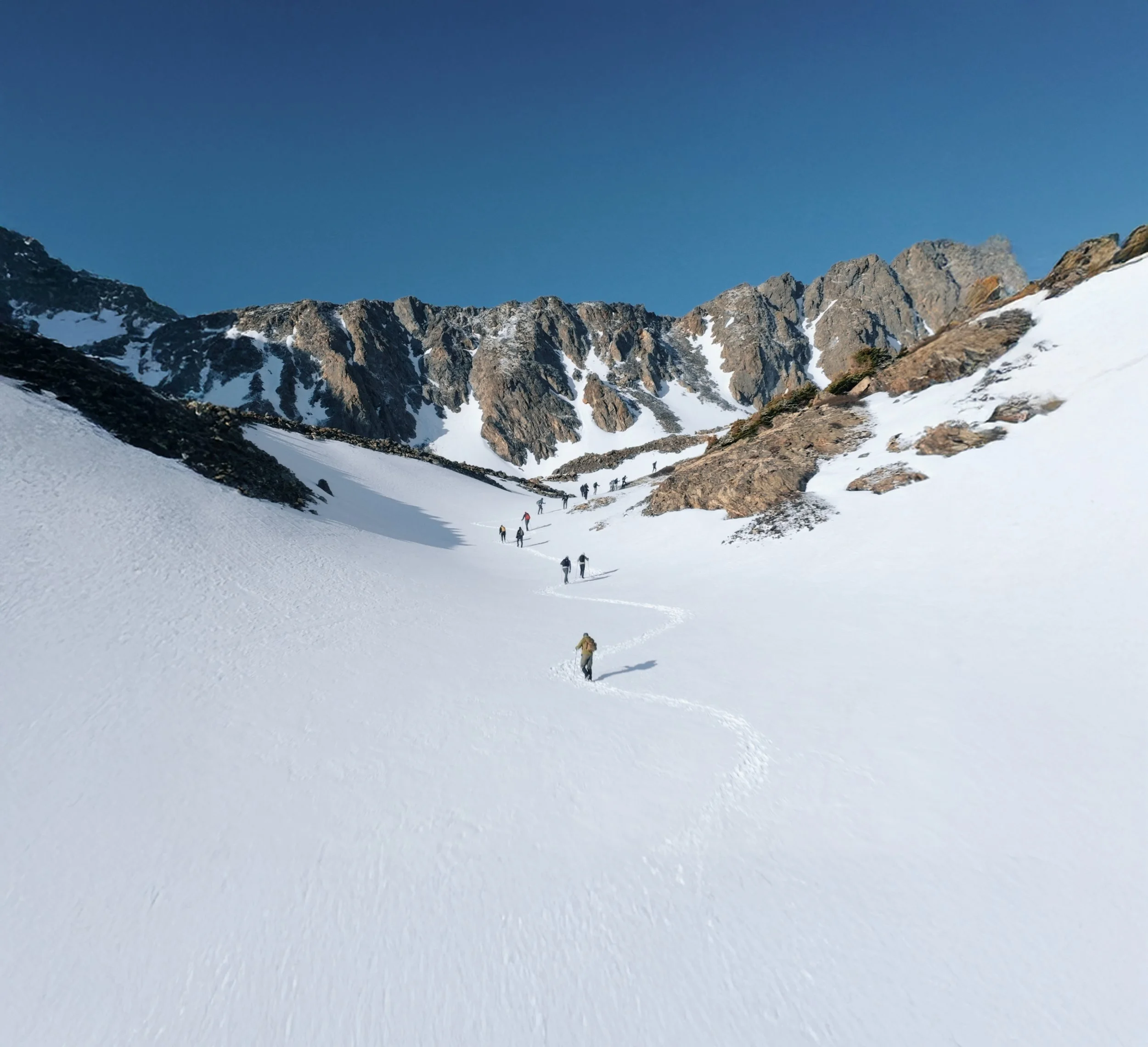 Group of hikers walking through snowy mountain landscape with rocky peaks and clear blue sky.