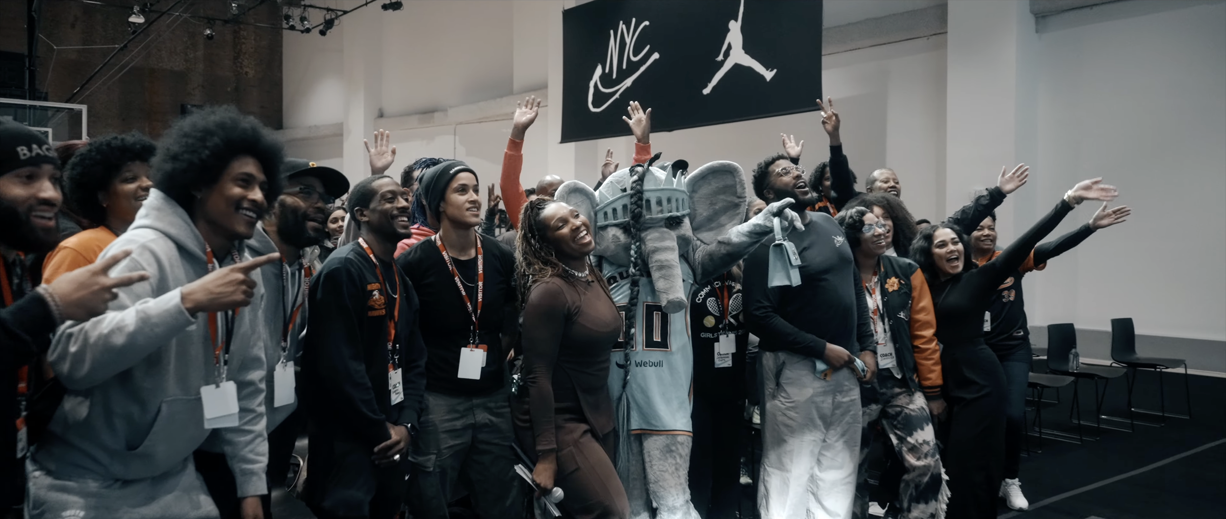 A group of diverse people at an indoor event, smiling and raising their hands, with a large sign showing the NYC and Jordan logos behind them.