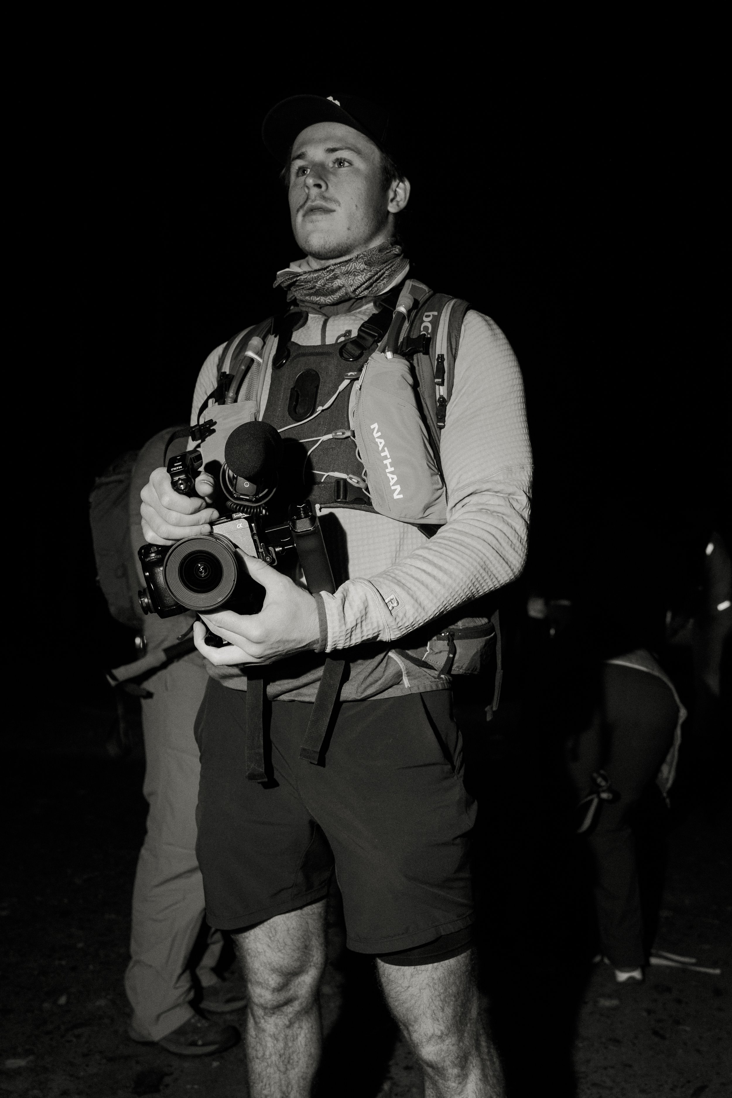 A man in outdoor gear holding a camera, standing outdoors at night.