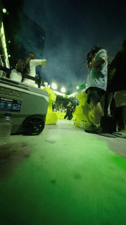 People at an outdoor event at night, with yellow barriers and bright lighting, some taking photos or videos, with a person walking in the background.