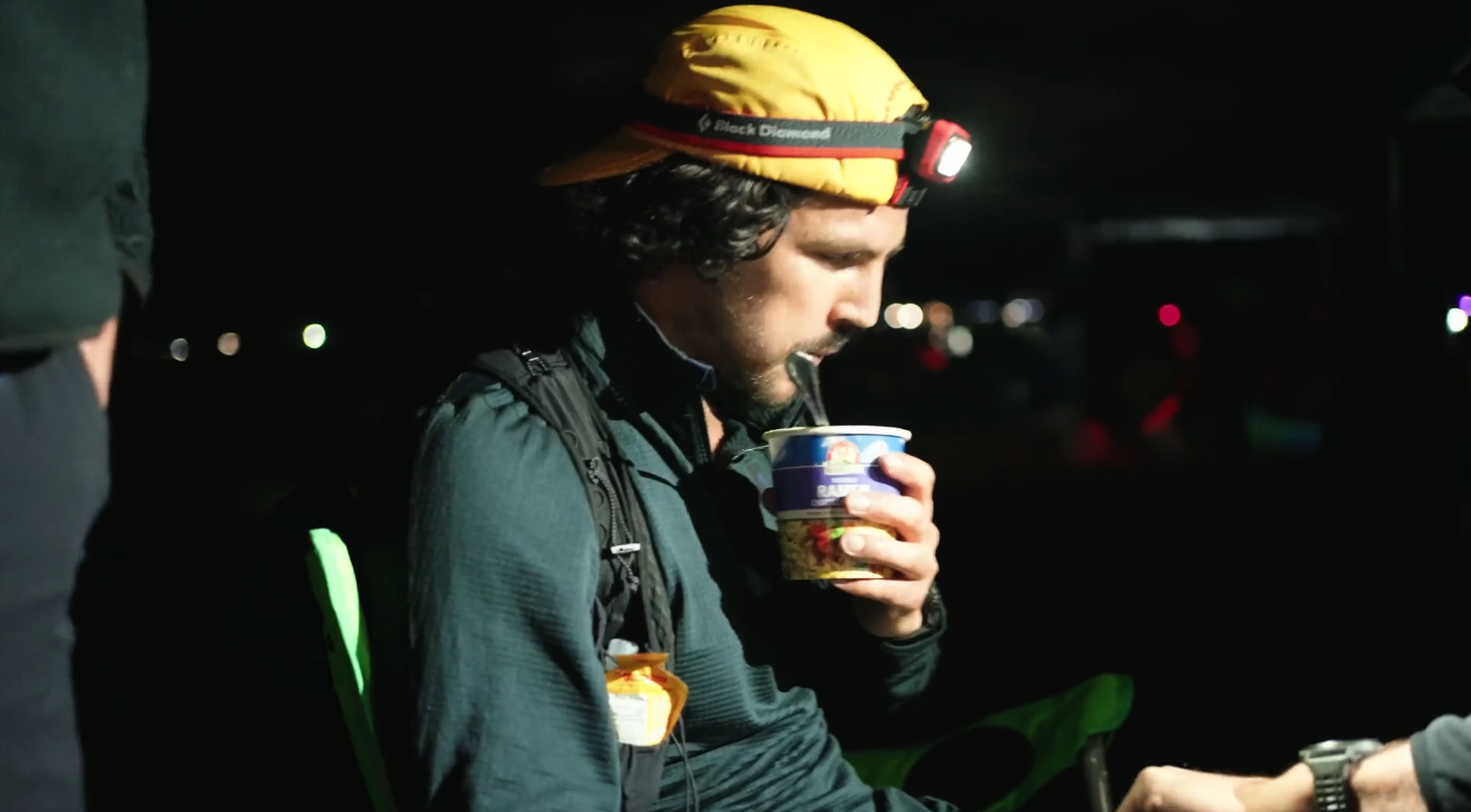 Man wearing a yellow headlamp and a yellow cap, with dark hair and stubble, drinking from a cup at night, dressed in dark outdoor clothing, seated outdoors with a green camping chair, illuminated by a headlamp against a dark background with small distant lights.