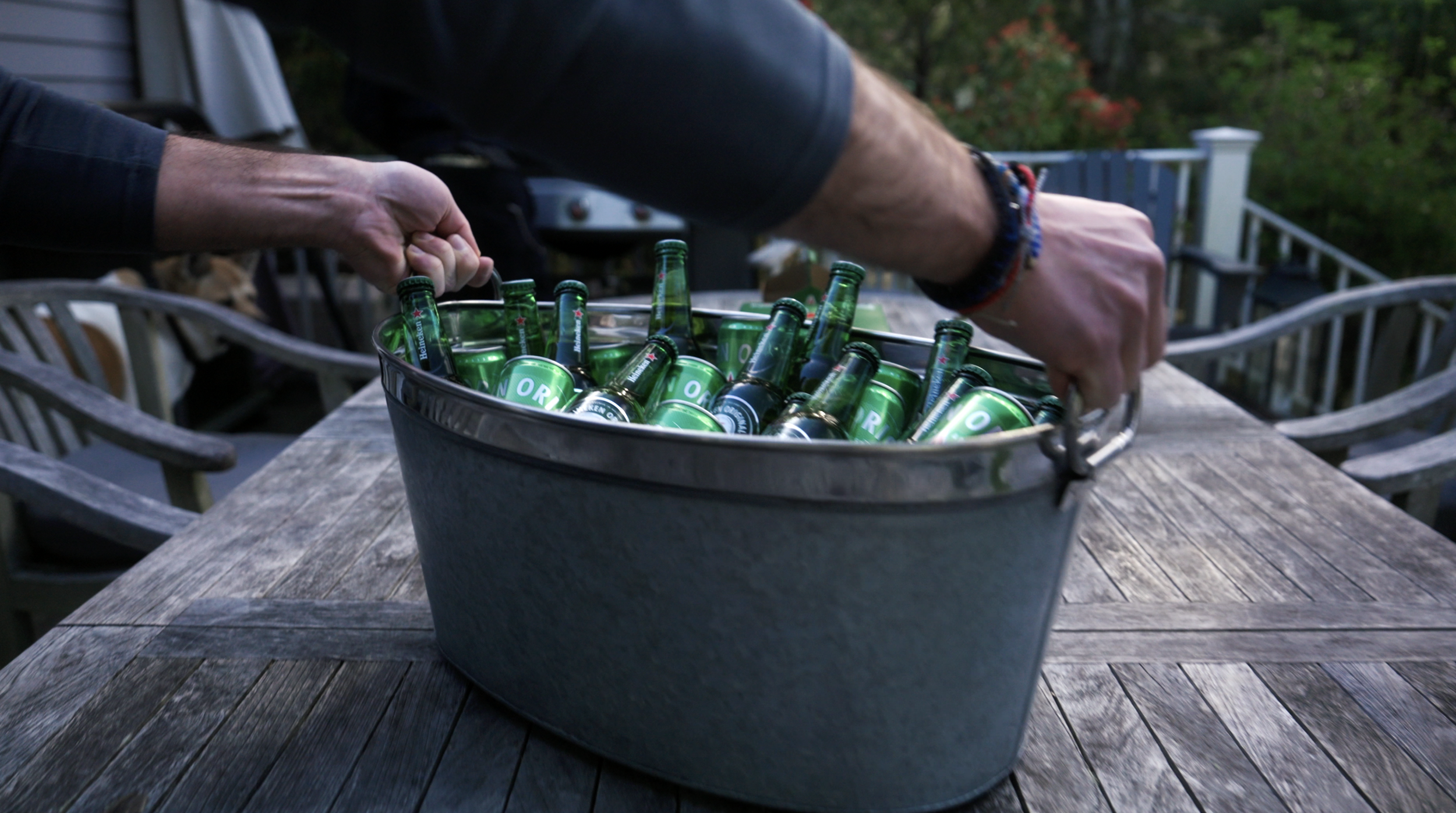 Person reaching into a metal bucket filled with Heineken and other beer bottles and cans, placed on a wooden outdoor table with outdoor chairs in the background.