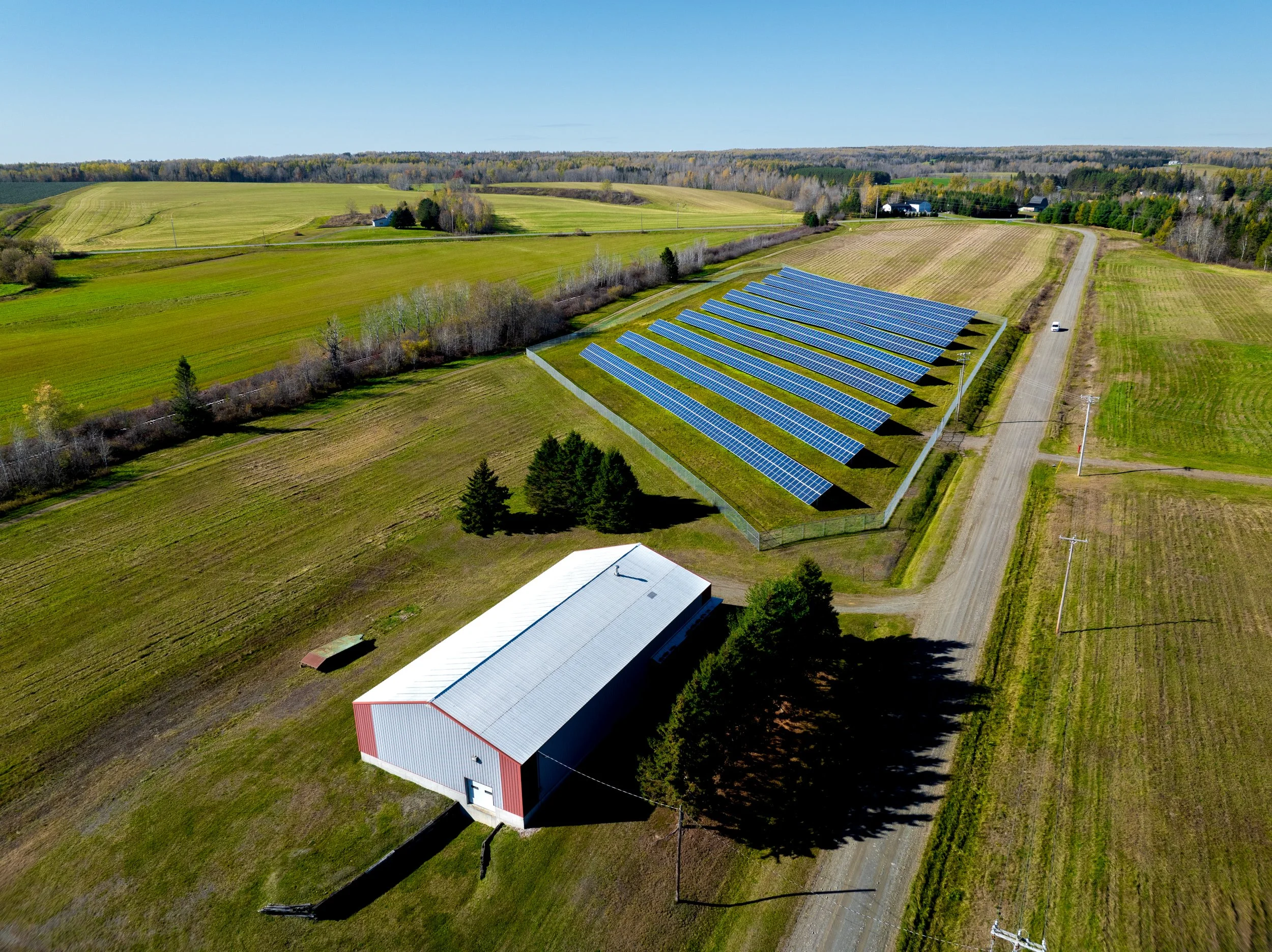 Aerial view of a rural farm with solar panels, a farming barn, and fields under blue sky.