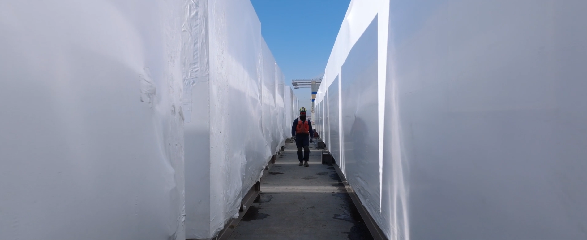 A person wearing a safety helmet, face mask, and orange safety vest walking between two large walls of ice.