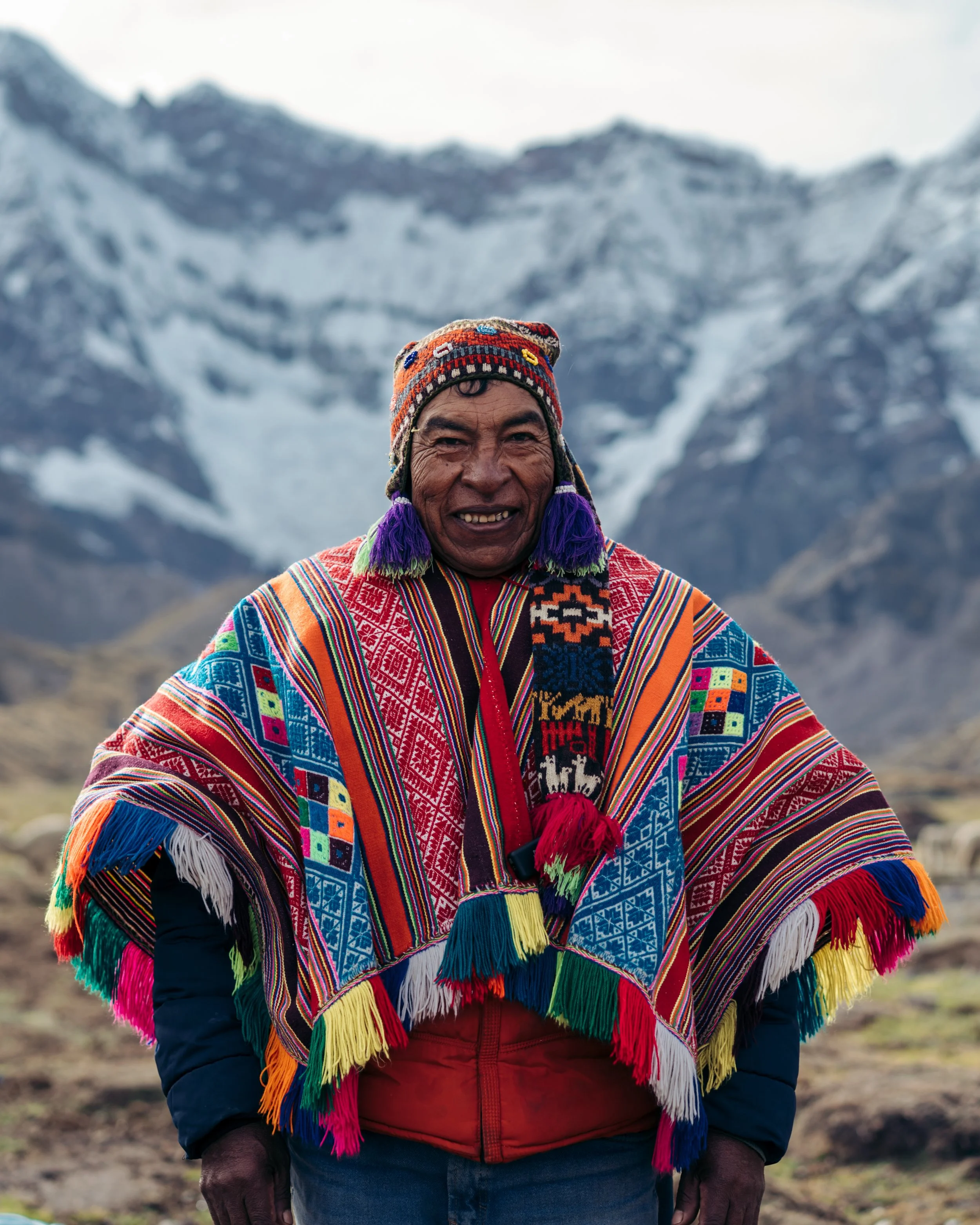 A smiling man wearing a colorful traditional Andean poncho and hat standing outdoors with snow-capped mountains in the background.