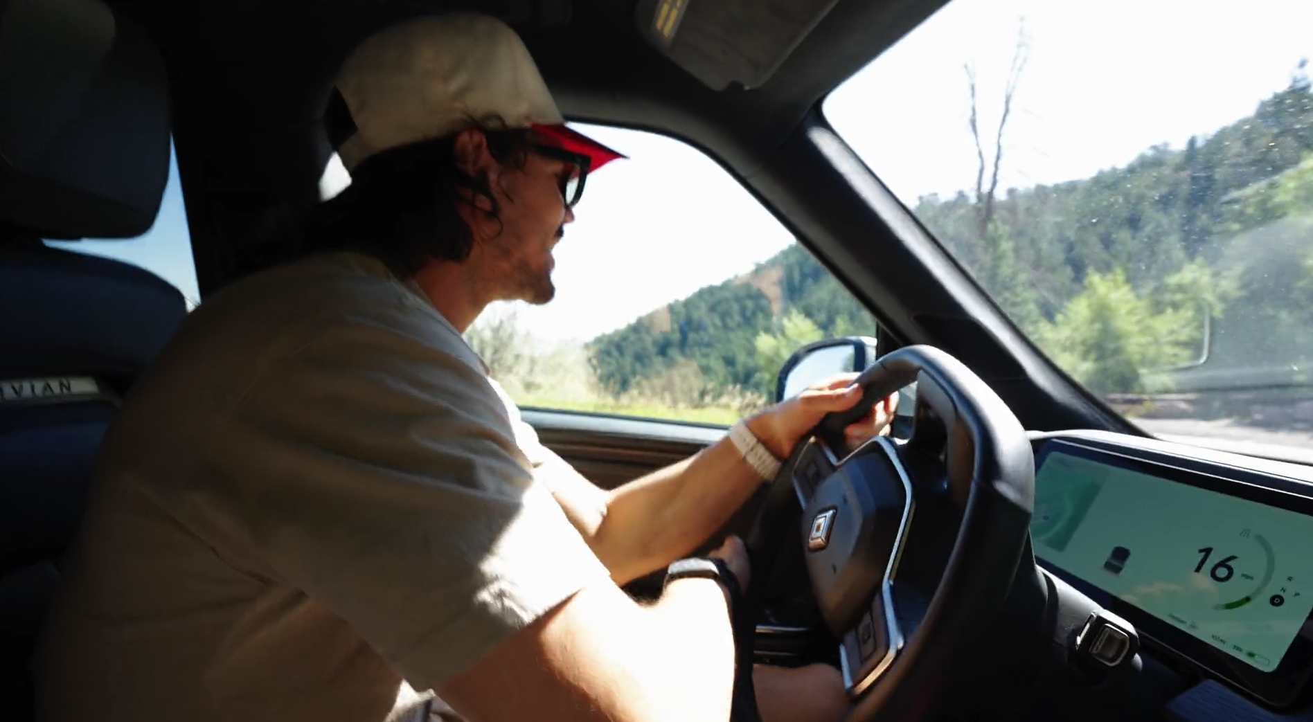 A man driving a vehicle on a scenic road with mountains and trees outside the window.