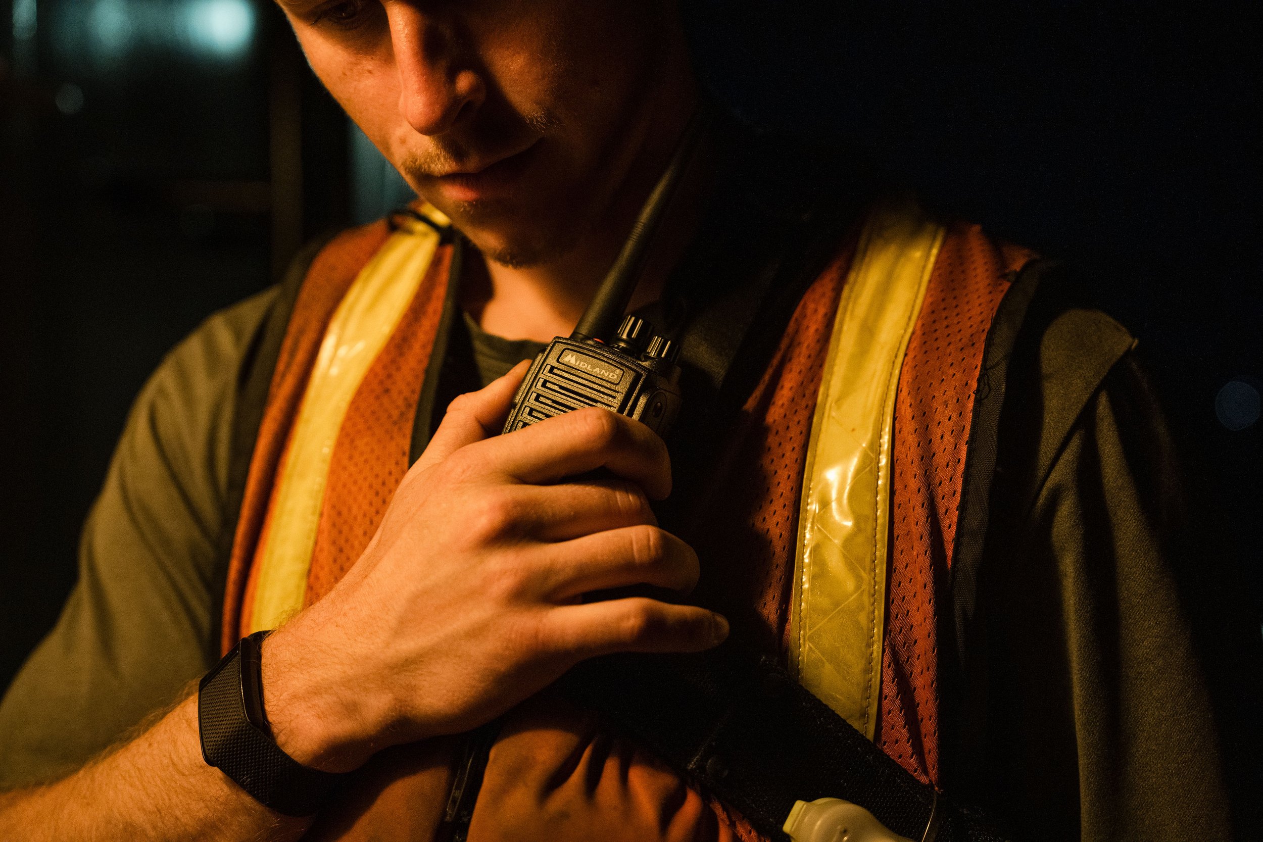 Man holding a walkie-talkie, wearing a safety vest, with a dark background.