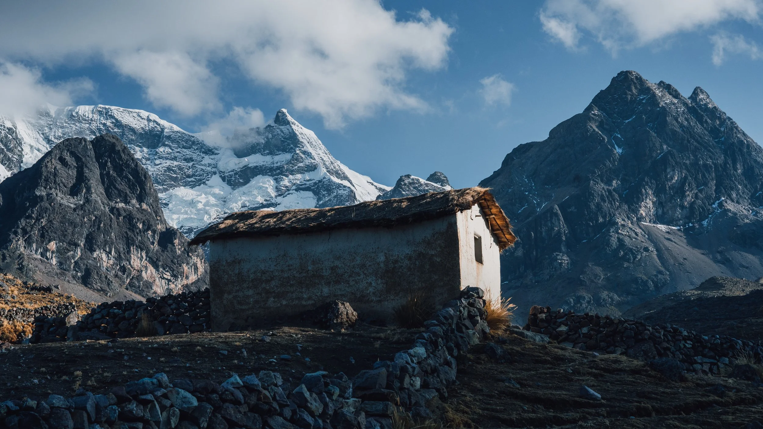 A small white hut with a thatched roof in a mountainous landscape with snow-capped peaks and a blue sky.