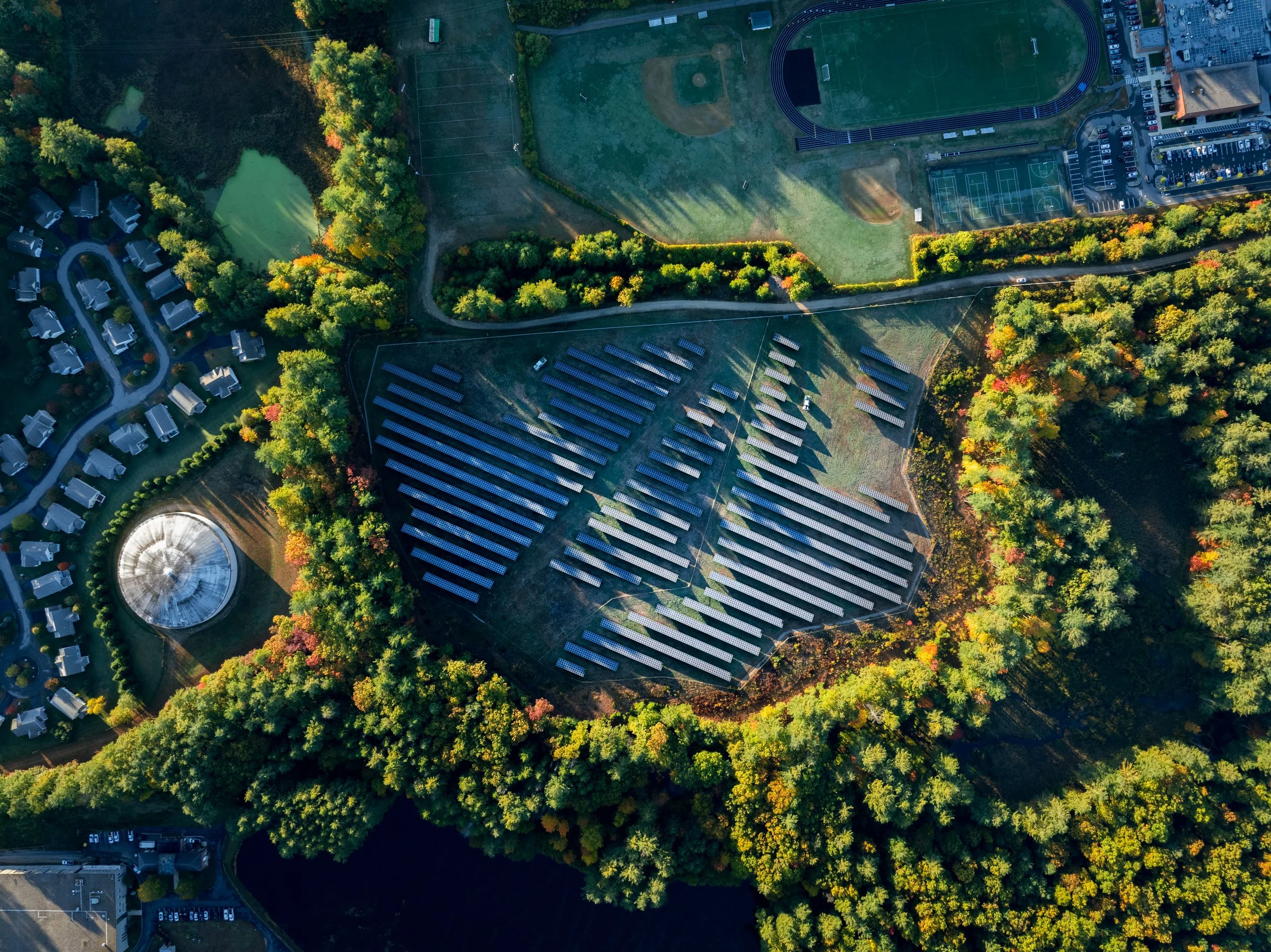 An aerial view of a park with a mix of green trees, adjacent sports fields, and a solar panel installation at the center.