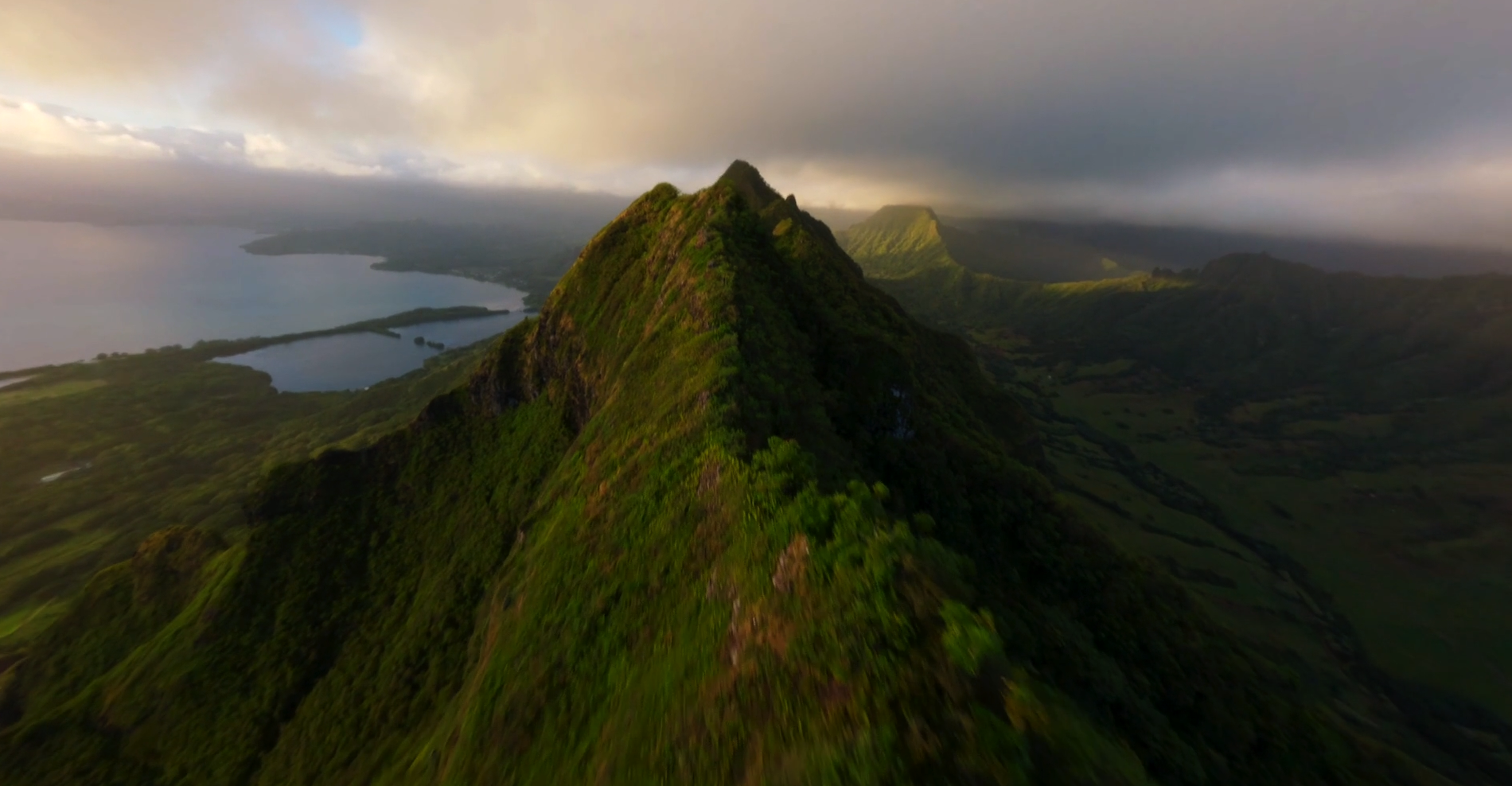 Aerial view of lush green mountains with a prominent peak, overlooking a body of water with islands, under partly cloudy skies.