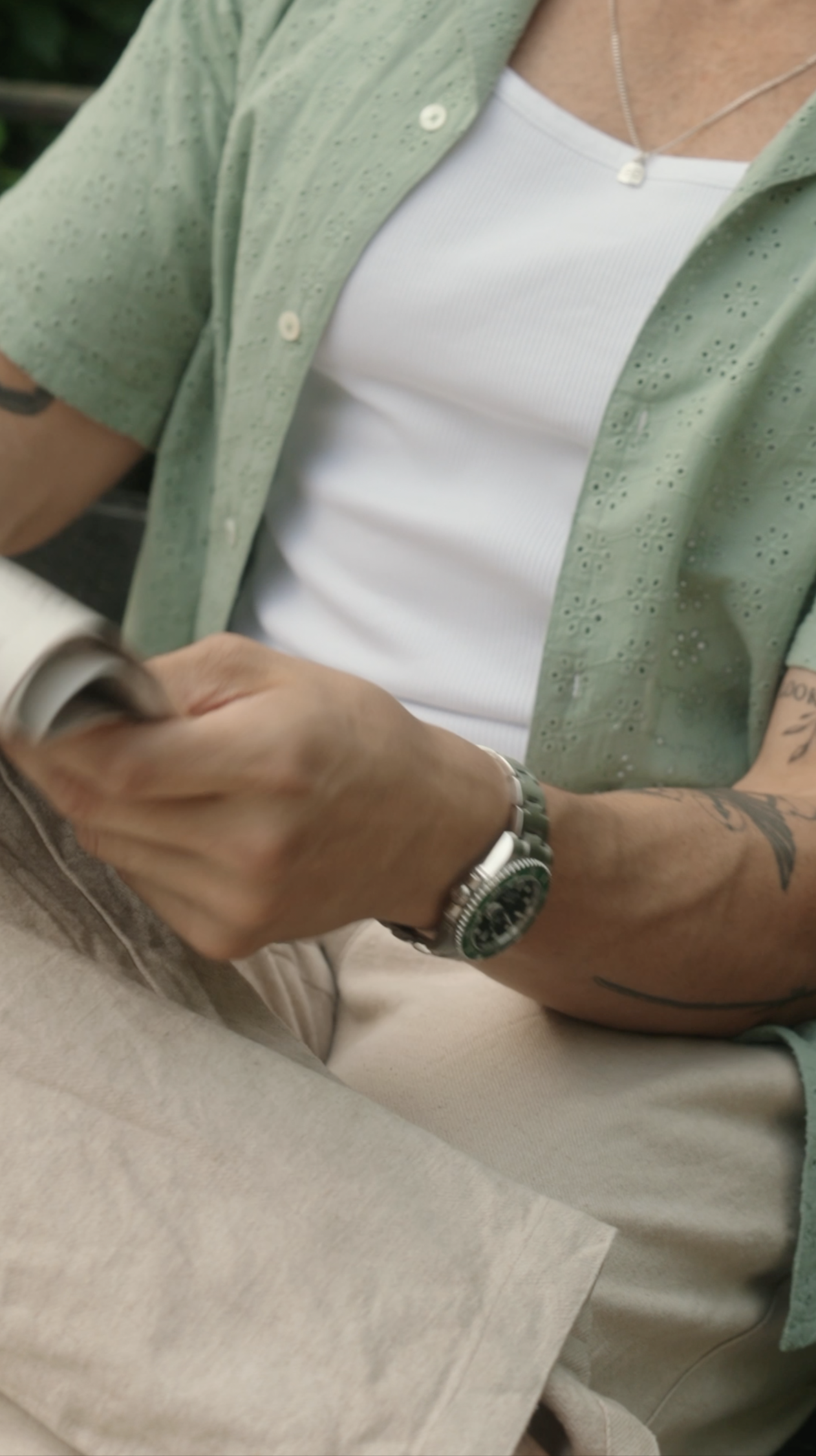 A man with tattoos on his arm, wearing a watch and a green shirt over a white t-shirt, sitting and holding a mug in one hand.
